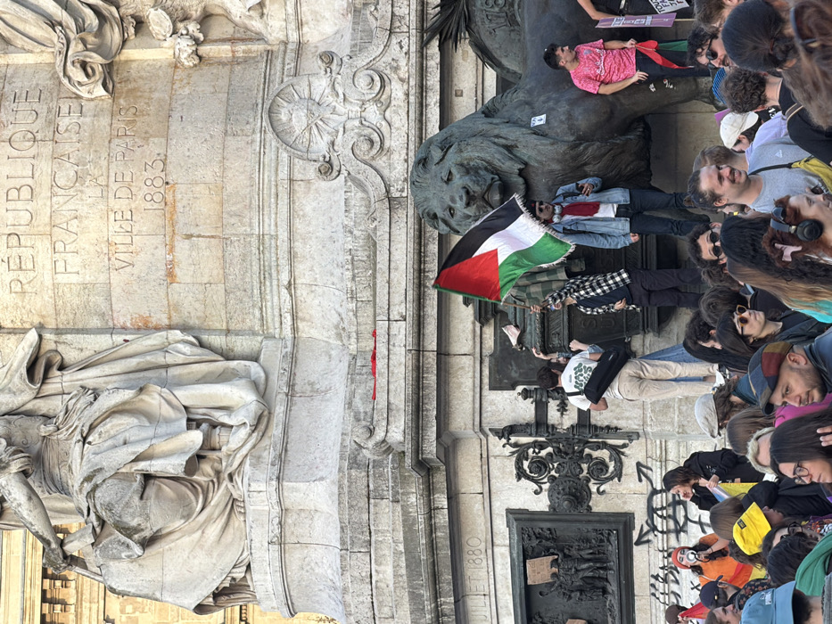 The scene depicts a gathering of individuals around the base of the Monument à la République in Paris, France.
The upper visible section of the monument consists of light-colored stone with a large allegorical statue, likely Marianne, seated and draped, partially visible at the top left. Below this, the monument's façade bears the carved inscription "RÉPUBLIQUE FRANÇAISE LA VILLE DE PARIS 1883." Further down on the left, "JUILLET 1880" is inscribed. The base features ornate bronze panels and a large bronze lion statue positioned on the right, facing right. A small, red fabric item lies on a stone ledge of the monument's base, near the center. Graffiti is present on the lower stone.
A crowd of diverse individuals occupies the foreground and midground. Several people are positioned on the lower tiers of the monument and on the bronze lion statue.
* Centrally, an individual wearing a plaid shirt and dark pants stands on the stone base of the lion statue, holding a Palestinian flag (black, white, green horizontal stripes with a red triangle at the hoist) aloft with both hands.
* To the left of the flag-holder, another person in a white t-shirt and light pants, carrying a black cylindrical bag, stands on a lower stone ledge, looking towards the crowd.
* To the right of the flag-holder, an individual in a denim jacket, red scarf, and sunglasses stands on the lion's back, actively viewing content on a mobile phone.
* Further right, a person in a pink patterned t-shirt stands on the lion's back, holding a smaller flag, partially visible, consistent with Palestinian colors.
* In the lower left foreground, a woman with orange hair and an orange mask holds a megaphone, appearing to address individuals to her left.
* The surrounding crowd is densely packed, with individuals of various ages and attire, many observing the monument or interacting with each other. Some individuals are smiling.
The environment is outdoors, under clear daytime lighting conditions. The monument exhibits some weathering and discoloration on its stone surfaces.