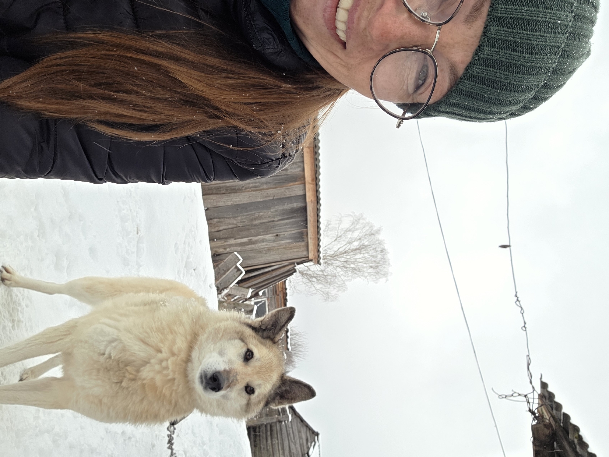 A woman in a green knitted cap and glasses smiles down at a light-colored, medium-sized dog with pointy ears standing in snow. The dog is chained. Behind the dog and woman are wooden structures, possibly buildings, and a bare tree covered in frost. The sky is overcast. The location is Зипуново, Russia.