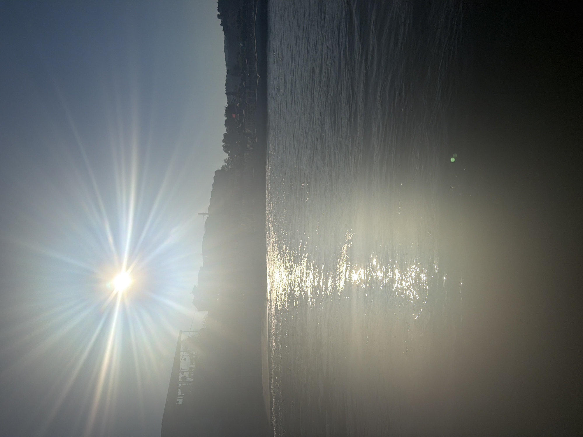 The image depicts a bright, sunlit scene over a body of water, likely the sea or a large lake, with a shoreline and buildings in the background. The sun is a prominent feature, positioned in the upper left quadrant, emitting strong rays of light that cause lens flare and illuminate the water with a shimmering reflection. The water itself is dark and appears slightly rippled. Along the shoreline, there are silhouetted structures, possibly buildings or cafes, suggesting a coastal town or resort. In the distance, hills or mountains can be seen, with more buildings and vegetation on their slopes. The sky is a clear, pale blue. The location is identified as Georgioupoli, Greece, a coastal town on the island of Crete known for its beaches and the freshwater lake of Kournas. The time of day appears to be morning or late afternoon, given the angle of the sun. There are no visible people or specific actions taking place. The overall impression is one of a serene and bright summer day.