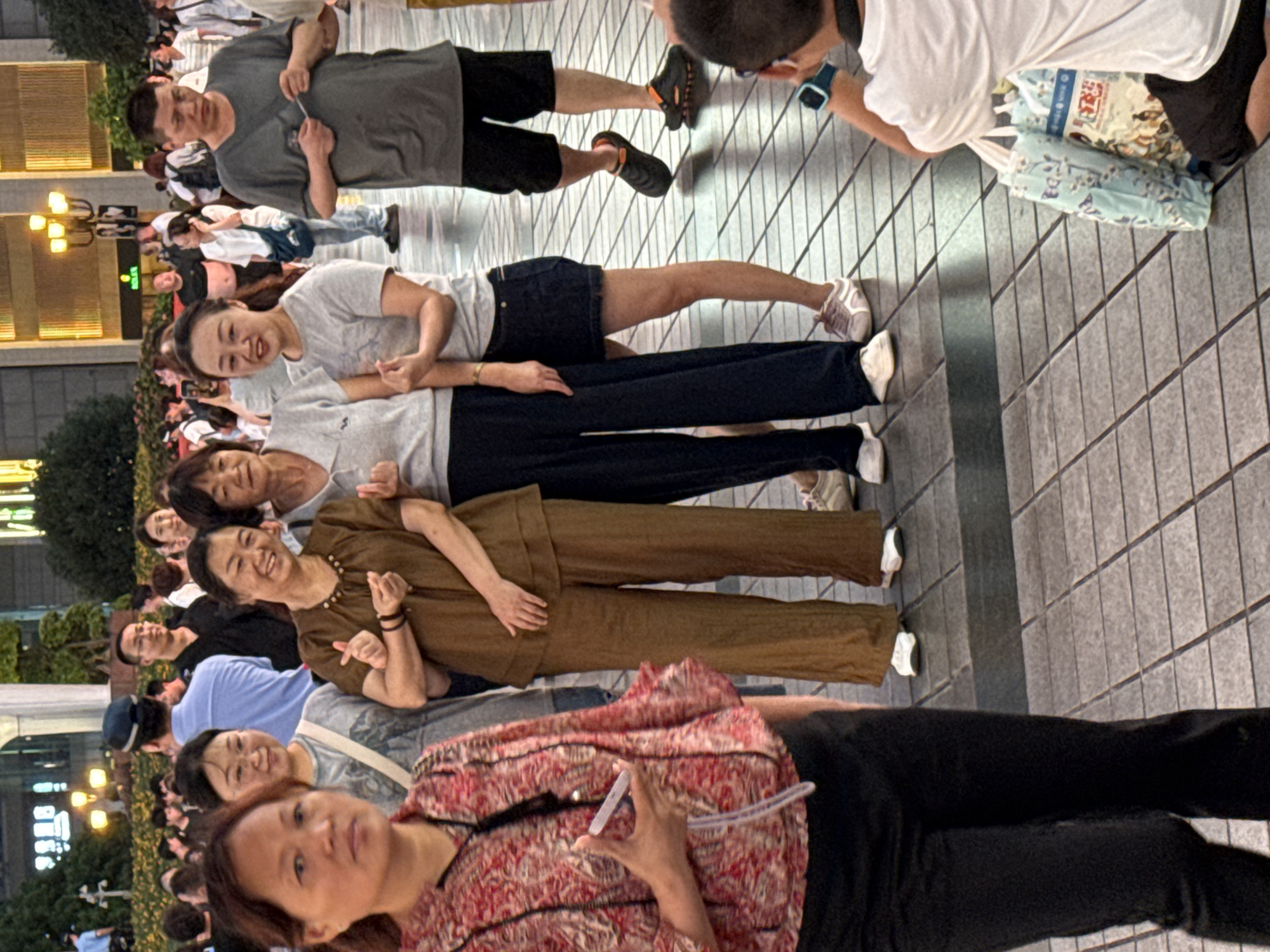 In Yuzhong District, China, a group of people are gathered outdoors on a tiled pedestrian walkway. The scene appears to be a public area, possibly a commercial district, given the illuminated buildings in the background.
In the foreground, a woman in a red and white patterned shirt and black pants is on the left, looking towards the camera. To her right, three women are standing together, appearing to be taking a photo together. The woman in the center is wearing a brown outfit, and the woman to her right is in a grey t-shirt and a dark blue skirt. The woman to the left of the brown-clad woman is wearing a grey t-shirt and dark pants.
Further back, a man in a grey t-shirt and black shorts is visible, holding a small item in his hand. He is looking down. Another man in a light blue shirt and dark cap is standing behind him. Other individuals and blurred figures are dispersed throughout the background.
The ground is paved with square grey tiles. Shopping bags are visible near the bottom right corner of the image. The lighting suggests it is late afternoon or early evening.