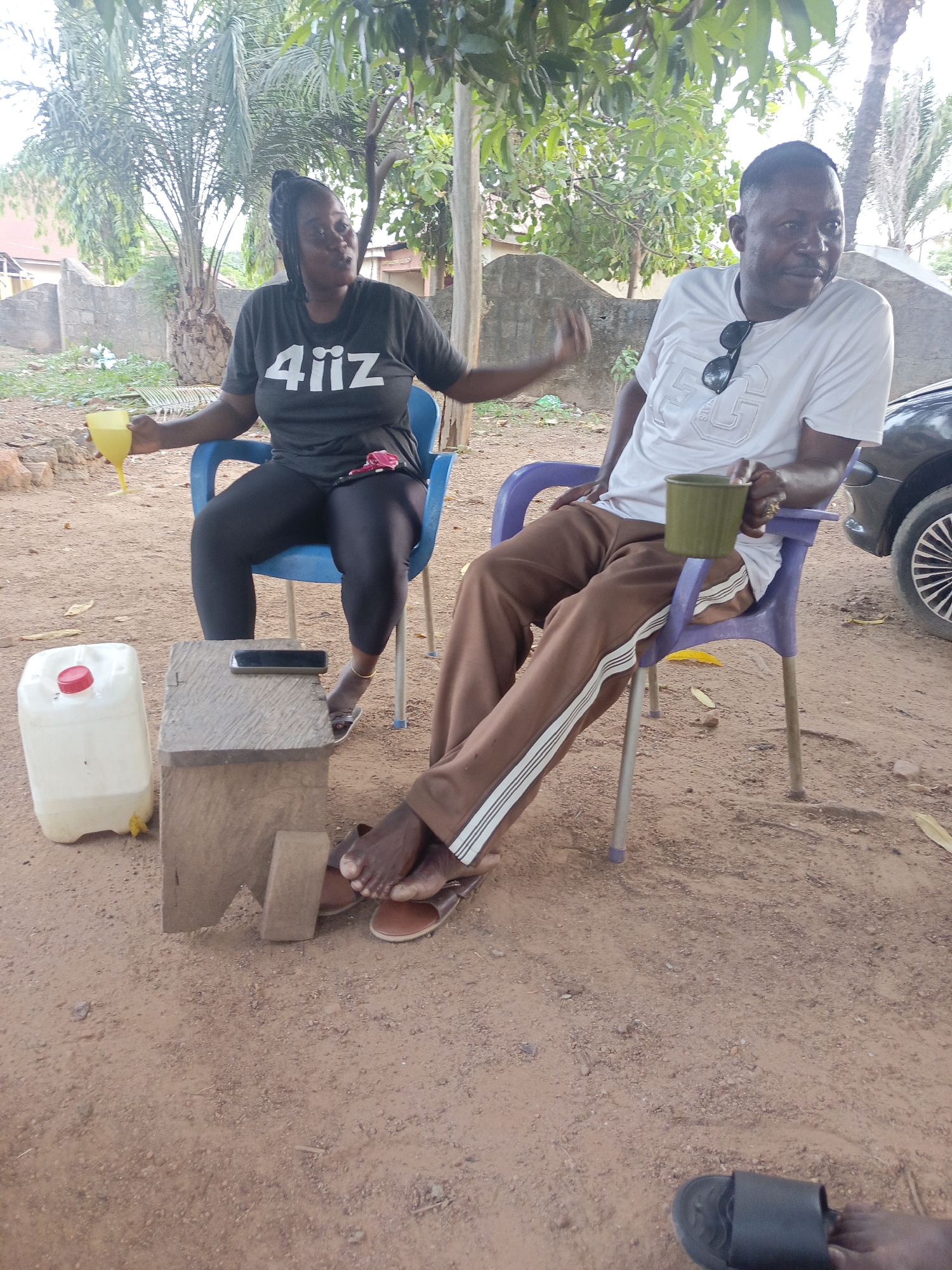 Two adults, a woman and a man, are seated outdoors on plastic chairs, engaged in conversation. The woman, positioned on the left in a blue chair, has dark skin and braided hair tied back. She wears a dark grey t-shirt, black leggings, and sandals. She holds a yellow-stemmed glass containing a light yellow beverage and gestures with her right hand. The man, on the right in a purple chair, also has dark skin and short dark hair. He wears a white t-shirt, brown track pants with white stripes, and brown sandals. Sunglasses are hung on his t-shirt. He holds a dark green mug and looks towards his right with a calm expression.
Between them, on the reddish-brown dirt ground, is a small, rustic wooden stool. A black smartphone rests on the stool. Next to the stool, a translucent white plastic jerrycan with a red cap and a yellow spigot sits on the ground. The setting is outdoors, likely a yard or informal gathering space, characterized by the dirt ground and lush green foliage from various trees, including palm trees, providing shade in the background. A low, weathered concrete block wall is visible behind the woman. The front part of a dark-colored car is partially visible on the far right. A black slipper is seen in the bottom right corner of the frame.
The overall atmosphere suggests a relaxed daytime gathering in a warm climate, consistent with the Akwanga, Nigeria context. The woman's t-shirt has the text "4iiZ" printed in white. The man's white t-shirt features a partial design that appears to include "5G".