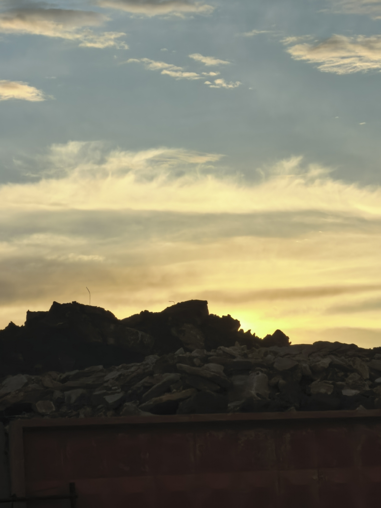 This is a vertical, medium shot photograph of a sunset over a pile of debris in an urban area. The sky is filled with wispy clouds, transitioning from pale blue at the top to bright yellow and orange near the horizon. A silhouette of jagged debris, possibly from a demolition site, rises against the sunset. A thin, dark pole, possibly a street lamp, is visible on the left side of the debris pile. The bottom portion of the image shows the top edge of a dark red, weathered container or dumpster. The overall scene suggests an industrial or construction area at dusk. The location is Unknown, Unknown.