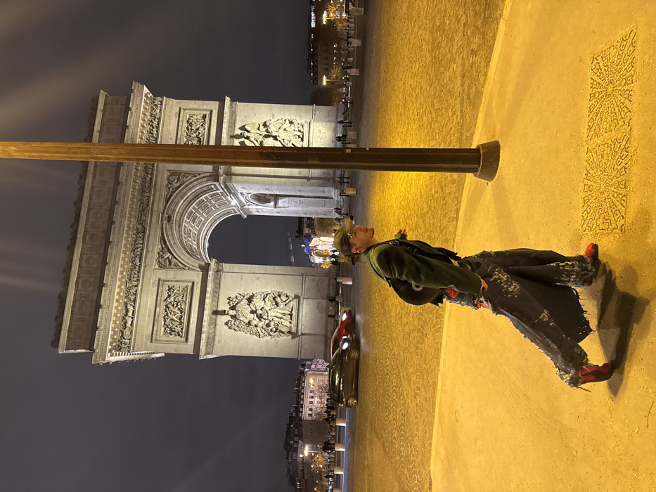 An outdoor nighttime scene in Paris, France, features the brightly illuminated Arc de Triomphe de l'Étoile centrally in the background, showcasing its intricate bas-reliefs and grand arches against a dark sky. In the immediate foreground, a person walks from left to right across a warmly lit, paved surface, angled slightly away from the viewer. This individual wears a dark baseball cap, dark green jacket, red undergarment, heavily distressed and wide-legged denim trousers with red textile accents and significant fraying, and bright red shoes. A black backpack is worn on their left shoulder. A tall, dark lamppost stands vertically in the mid-ground to the right, illuminated by a warm yellow-orange light that also bathes the foreground pavement. The paved ground exhibits a textured pattern in the lower right corner. In the middle distance, on the traffic roundabout surrounding the Arc de Triomphe, two blurred vehicles—one dark and one red—are in motion. Several indistinct figures are visible near the base of the monument. Distant Parisian buildings with illuminated windows are discernible in the far background.