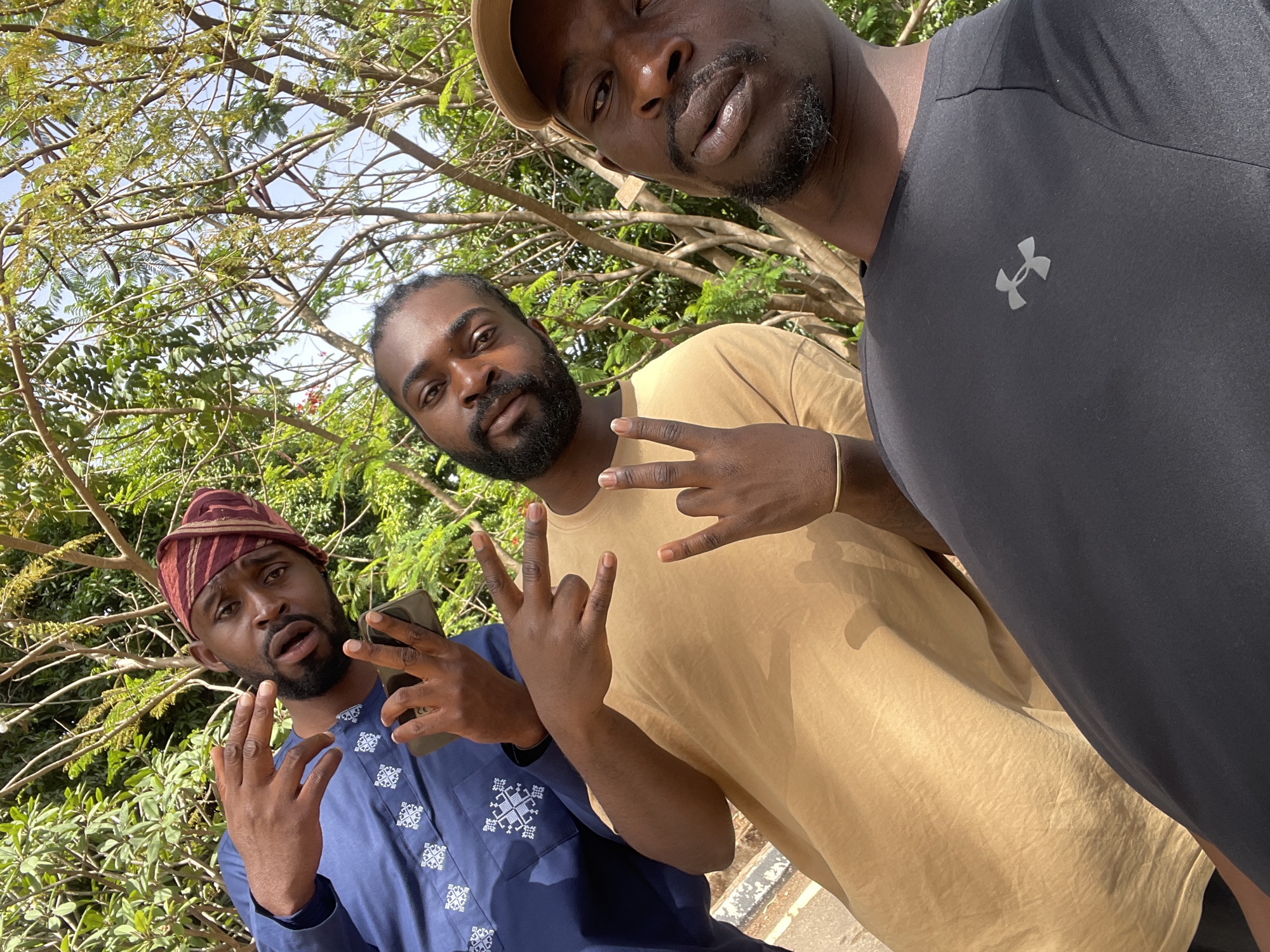 A bright outdoor selfie captures three men of African descent in what appears to be a casual, friendly moment, set against a backdrop of lush green trees and a clear blue sky, indicative of a sunny day in Jos, Nigeria.
On the left, one man is clad in a traditional multi-colored striped cap, possibly a gèlè or similar Yoruba headwear, and a blue garment featuring distinctive white embroidered patterns, suggesting a dashiki or traditional attire. He has a full beard, holds a dark smartphone in his left hand, and makes a hand gesture with both hands, while his mouth is slightly open.
In the center, another man, sporting dreadlocks and a beard, wears a plain light brown t-shirt. He looks directly at the camera, making a "rock on" hand gesture with his right hand.
The man closest to the camera, presumably taking the selfie, is positioned on the right. He wears a dark grey t-shirt with the iconic Under Armour logo visible on the left chest and a light-colored baseball cap. He has a neatly trimmed beard or stubble.
The men appear relaxed and engaged, with their poses and hand gestures conveying a sense of camaraderie. The setting is clearly natural and vibrant, emphasizing the outdoor environment.