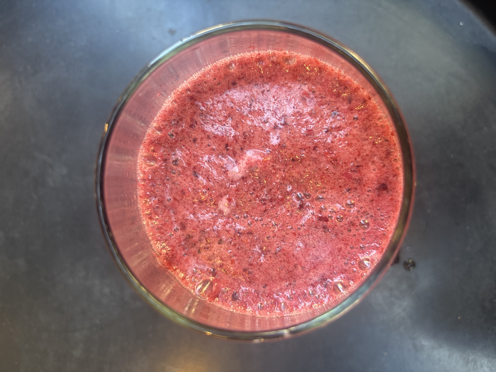 A top-down photograph captures a clear glass containing a vibrant reddish-pink liquid, indicative of a fruit smoothie or blended beverage. The surface of the drink is slightly frothy with numerous small bubbles, suggesting it was recently prepared. Darker specks, likely particles of fruit pulp or seeds, are visible throughout the liquid, contributing to its textured appearance. The glass rests on a dark, uniform surface, possibly a countertop or table, which provides a minimalist background. The surface appears non-reflective and offers no specific material identification beyond its dark hue. There are no people, explicit activities, or visible text in the image. The time of day, weather, and specific location cues within Buenos Aires, Argentina, are not discernible from the photograph itself. The image focuses entirely on the prepared drink.