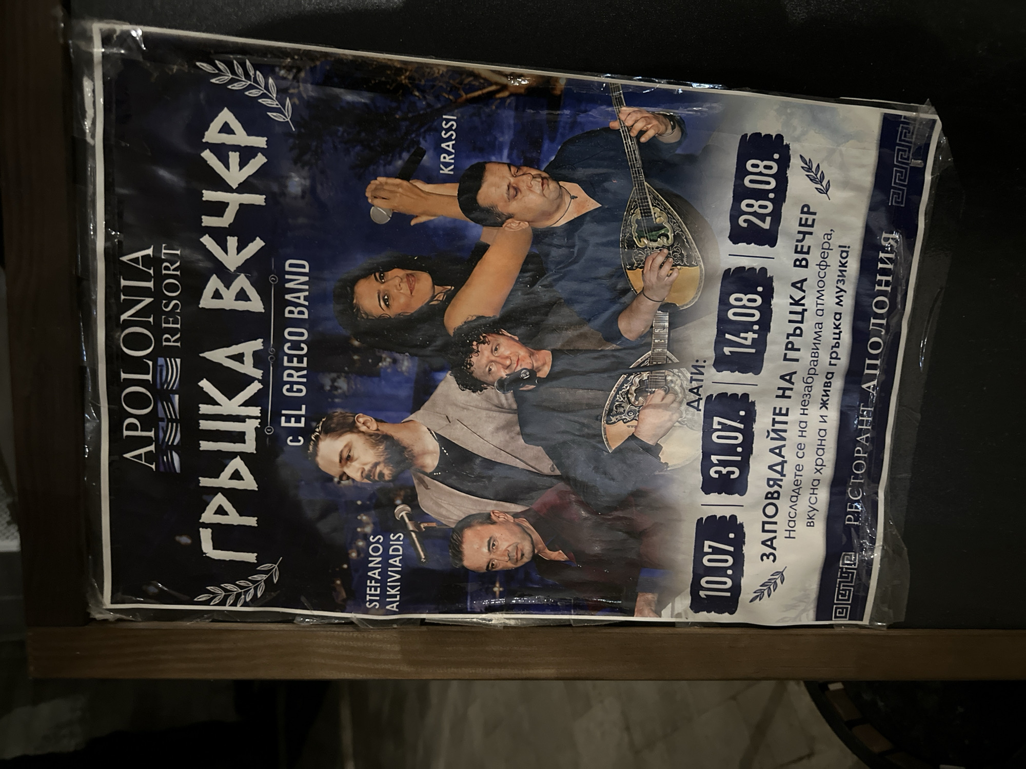 The image shows a poster advertising a "Greek Night" at the Apollonia Resort in Sozopol, Bulgaria. The poster features a group of musicians, with a female singer holding a microphone and several men playing traditional Greek instruments like the bouzouki. Text on the poster indicates dates for the event: July 10, July 31, August 14, and August 28. It also mentions "unforgettable atmosphere, delicious food and live Greek music." The poster appears to be laminated and is affixed to a wooden board, suggesting it's displayed outdoors or in a public space. The background of the poster depicts a dark, starry sky.