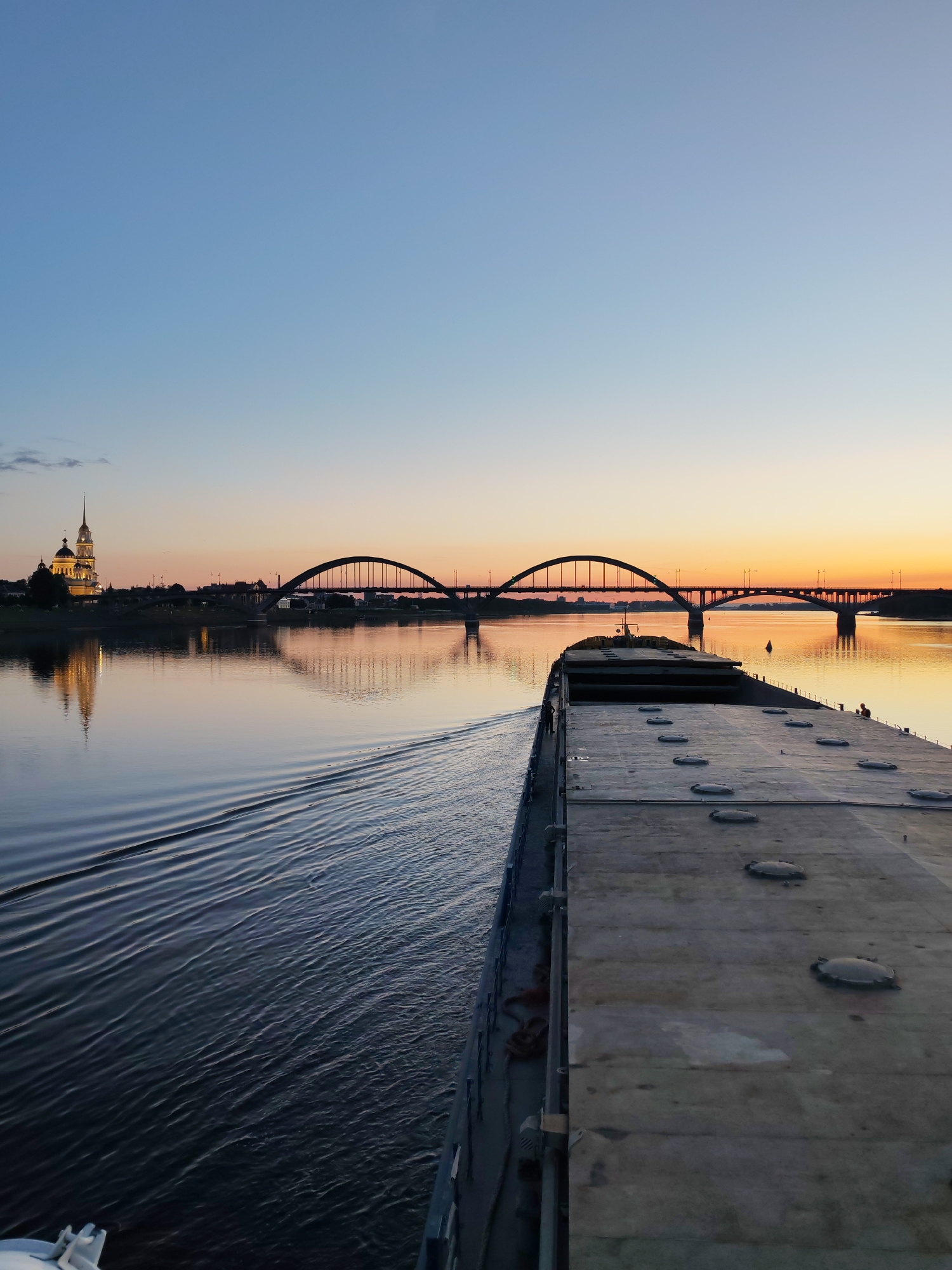 In Rybinsk, Russia, at sunset, a barge travels along a calm river. The barge, partially visible in the foreground, has a long, flat deck with multiple circular hatches. In the distance, a bridge with multiple arches spans the river. On the left bank, a church with a tall, slender steeple is illuminated, its reflection shimmering on the water. The sky displays a gradient of blue and orange hues. The water's surface is rippled, indicating the barge's movement.