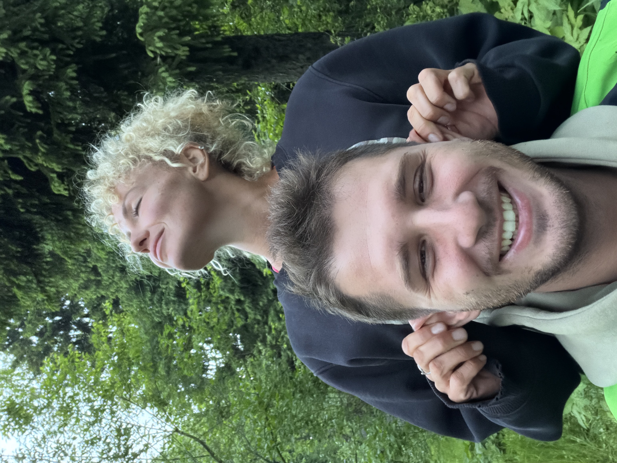 Two people are in the image. A woman with blonde, curly hair is standing behind a man with short brown hair and a beard. The woman's hands are positioned near the man's ears. The man is smiling. They are outdoors, with green foliage visible in the background. The specific location within the city of Trudovaya, Russia, is not discernible from the image.