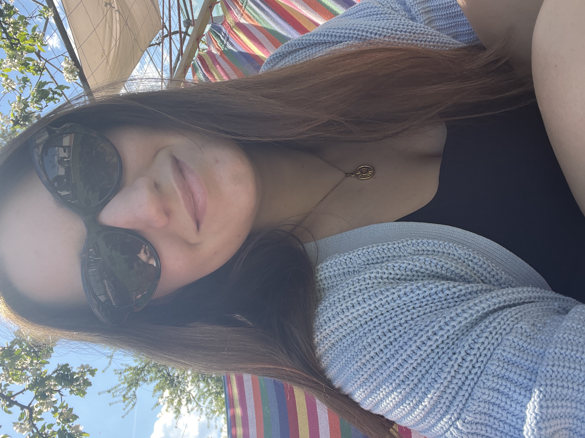 A medium shot shows a woman wearing sunglasses, a light blue cardigan, and a black top. She is wearing a gold necklace with a round pendant. She is smiling slightly. The background includes a striped hammock, trees with white blossoms, and a structure that resembles a partial sailboat or awning. The location appears to be outdoors.