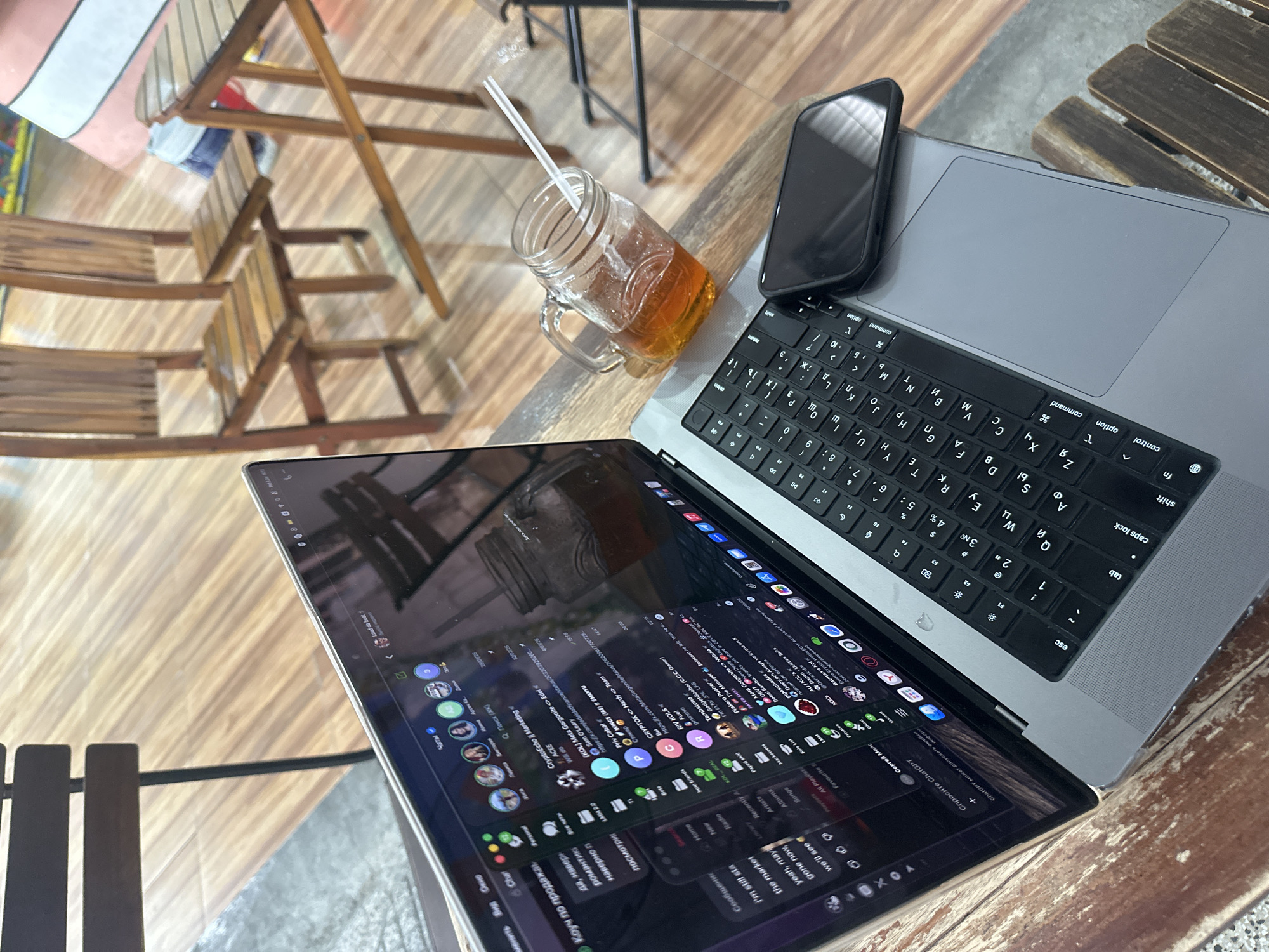 An overhead shot captures a workspace setup on a textured wooden table, likely in a cafe or co-working space in Nha Trang. A silver laptop, possibly a MacBook Pro, is open, displaying a screen full of chat applications (resembling Discord) and a music player, all in dark mode. The laptop's keyboard features both Latin and Cyrillic characters, indicating its use in a region where such languages are common. A black smartphone in a case rests on the laptop's palm rest, partially obscuring the trackpad. Next to the laptop, a glass jar mug with a handle contains an amber-colored liquid, likely iced tea, with a white straw.
The laptop screen shows numerous chat channels and messages. Notable text includes channel names like "CryptoEcho || Marketing," "PO WINGS (IAS) II SMANYU," and "KOL DEAL," along with user messages such as "I'm in for 5%, LFG," "yeah, may gone now, we'll see," and "порхай как бабочка" (fly like a butterfly). There are also references to "ChatGPT" and menu items like "Radio," "Artists," and "Albums." The background is out of focus but shows wooden folding chairs and parts of other tables, contributing to the casual cafe atmosphere. The overall scene suggests someone is engaged in online work or communication while enjoying a beverage.