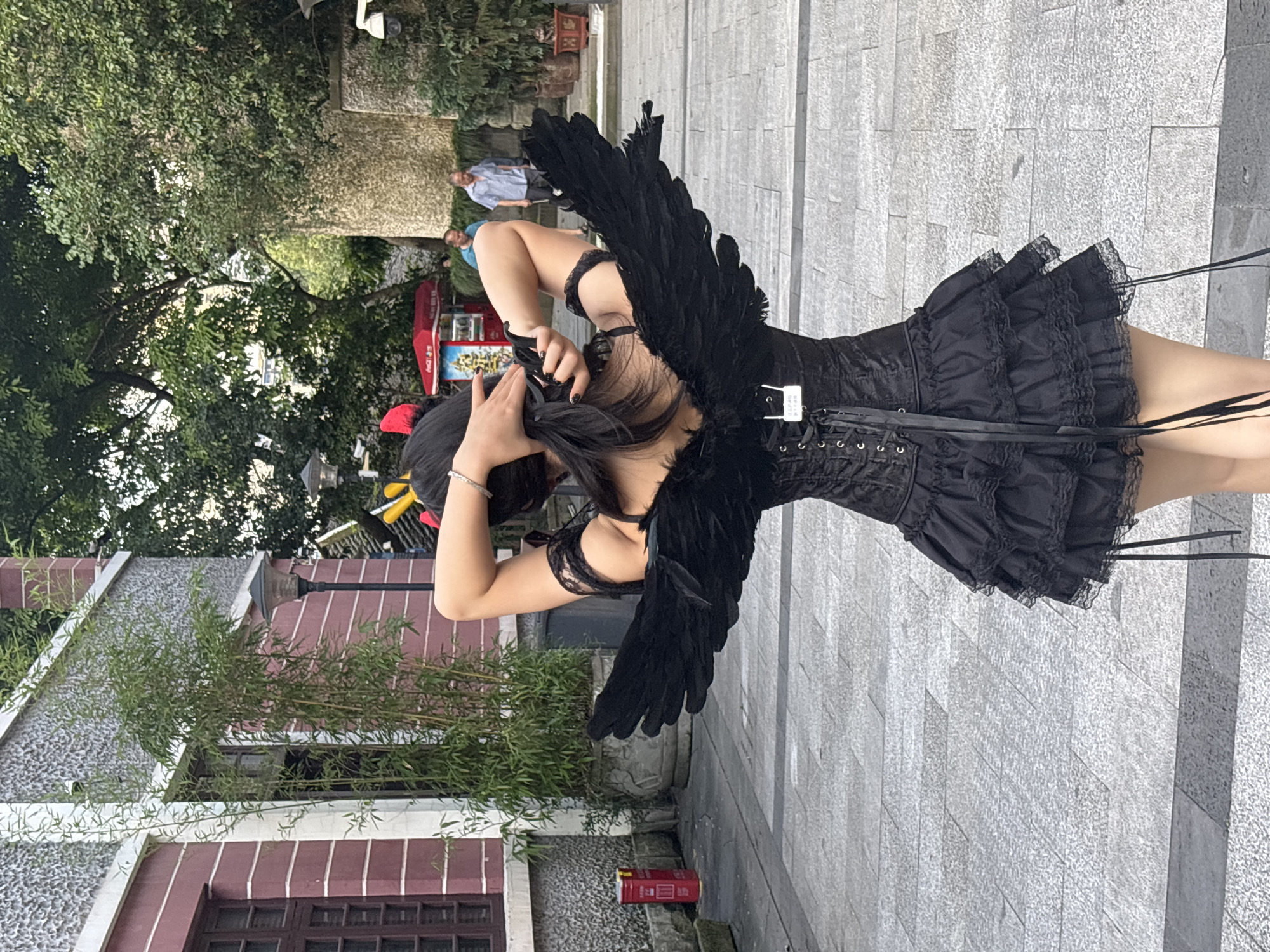 In Yuzhong District, China, a person wearing a black corset, a ruffled skirt, and black feather wings stands outdoors. The person has their hands behind their head, adjusting their hair. Two men are visible in the background, one in a blue shirt and the other in a light-colored shirt. A red kiosk with a sign featuring Chinese characters is also present. The ground is paved with gray stones. The architecture in the background includes a building with maroon and white siding and a textured stone wall. Green foliage and trees are scattered throughout the scene.
