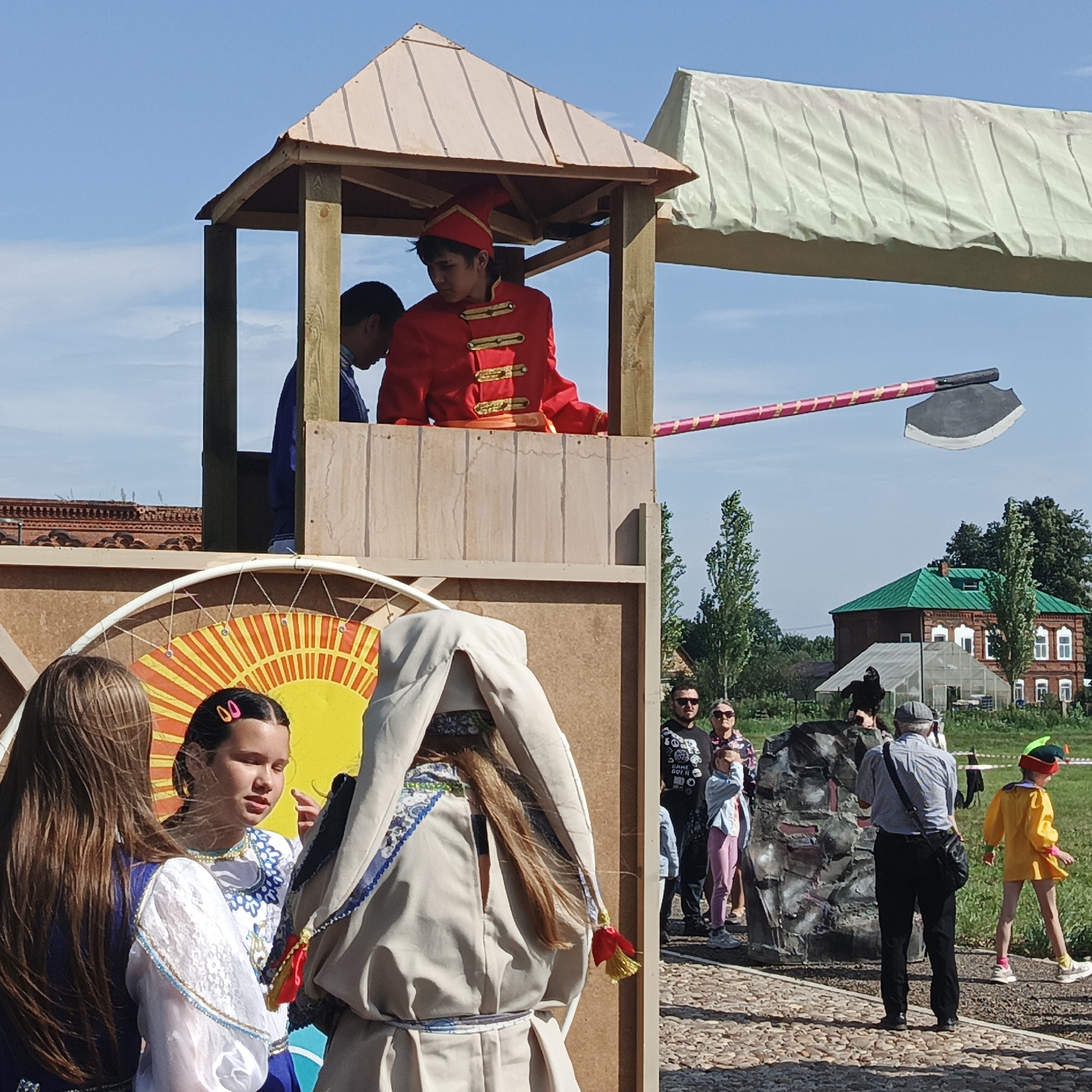 The image depicts an outdoor scene on a sunny day, likely at a cultural event or festival in Nikolo-Beryozovka, Russia. In the foreground, several individuals are gathered. Two young women are looking towards the left side of the frame, one wearing a blue and white traditional garment, the other with pink barrettes in her hair, also in traditional attire. A third person, also in traditional clothing, is facing away from the camera.
To the left, a wooden structure resembling a small watchtower is visible. Inside the tower, a young man in a red, gold-trimmed traditional costume and hat is looking down. Behind him, another person in a blue garment is partially visible.
A large, colorful circular decoration with a sun motif is mounted on the wooden structure. Extending from the watchtower towards the right, a long pole with a large, dark axe-head attached to one end is held horizontally in the air. The pole is decorated with a pink and yellow striped pattern.
In the background, to the right, several other people are observing the scene. Among them, a person in a yellow outfit with a green and red cap is walking away from the camera. Beyond the people and the event setup, a brick building with a green roof and several windows is visible, indicative of rural Russian architecture. Trees and a clear blue sky with some clouds complete the environment. The ground in the foreground is paved with small stones.
