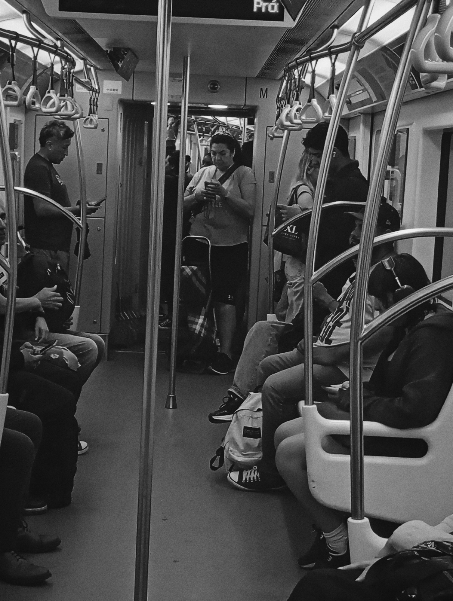 The scene depicts the interior of a Subte (subway) car in Buenos Aires, Argentina, presented in black and white. Approximately ten individuals are visible, occupying seats and standing in the aisle. Most passengers are engaged with their mobile phones, holding them at chest or waist level and looking downwards.
On the left, a man with short, dark hair stands, holding a phone in his right hand. Further back, another woman stands, also holding a phone. In the mid-ground, a woman with dark hair stands near a vertical pole, looking at her phone. She wears a light-colored top and dark shorts, with a patterned bag hanging from her shoulder. Her right foot, clad in a dark sneaker, is visible.
To the right, several passengers are seated. One individual on the rightmost seat wears a dark hooded garment and white socks with dark sneakers, also focused on a phone. Adjacent to them, another person with a dark hat and white t-shirt with a graphic design sits, looking straight ahead, with a backpack on the floor beside them. Further left, a person is seated, holding a dark bag on their lap. Above the seated passengers, a system of horizontal bars and numerous white hanging grab handles is visible. Vertical support poles run throughout the car's interior. In the upper background, overhead signage is partially visible, including a sign displaying "Pró." and another with the letter "M." The floor is dark and uniform. The lighting appears to be overhead fluorescent, casting soft shadows. No direct interactions between passengers are evident; the primary action is individual focus on personal devices.