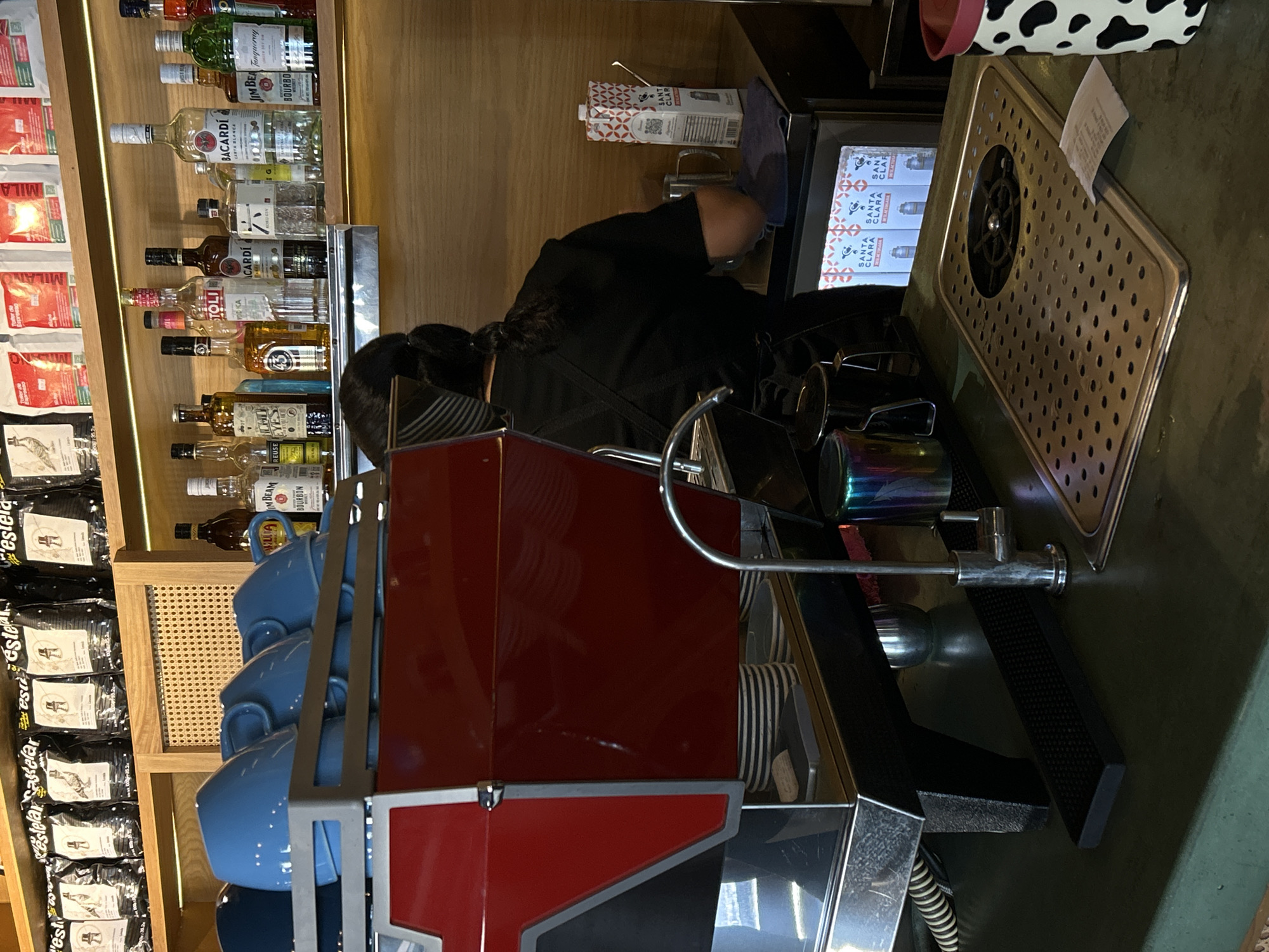 Here is a detailed description of the image:
The image shows a cafe or bar setting, potentially located in Mazatlán, Mexico, given the Spanish product labels. A person in a black uniform is working at an espresso machine. The machine is red and has a stack of blue coffee cups attached to its side. Behind the person and the espresso machine, there is a shelf with various bottles of alcohol, including brands like Bacardi and Jim Beam. There are also packages of coffee beans labeled "estelar" on the shelf. Cartons of "Santa Clara" milk are visible above the espresso machine. A metal grate with a cup rinser is located on the right. The general atmosphere suggests a commercial establishment preparing beverages.