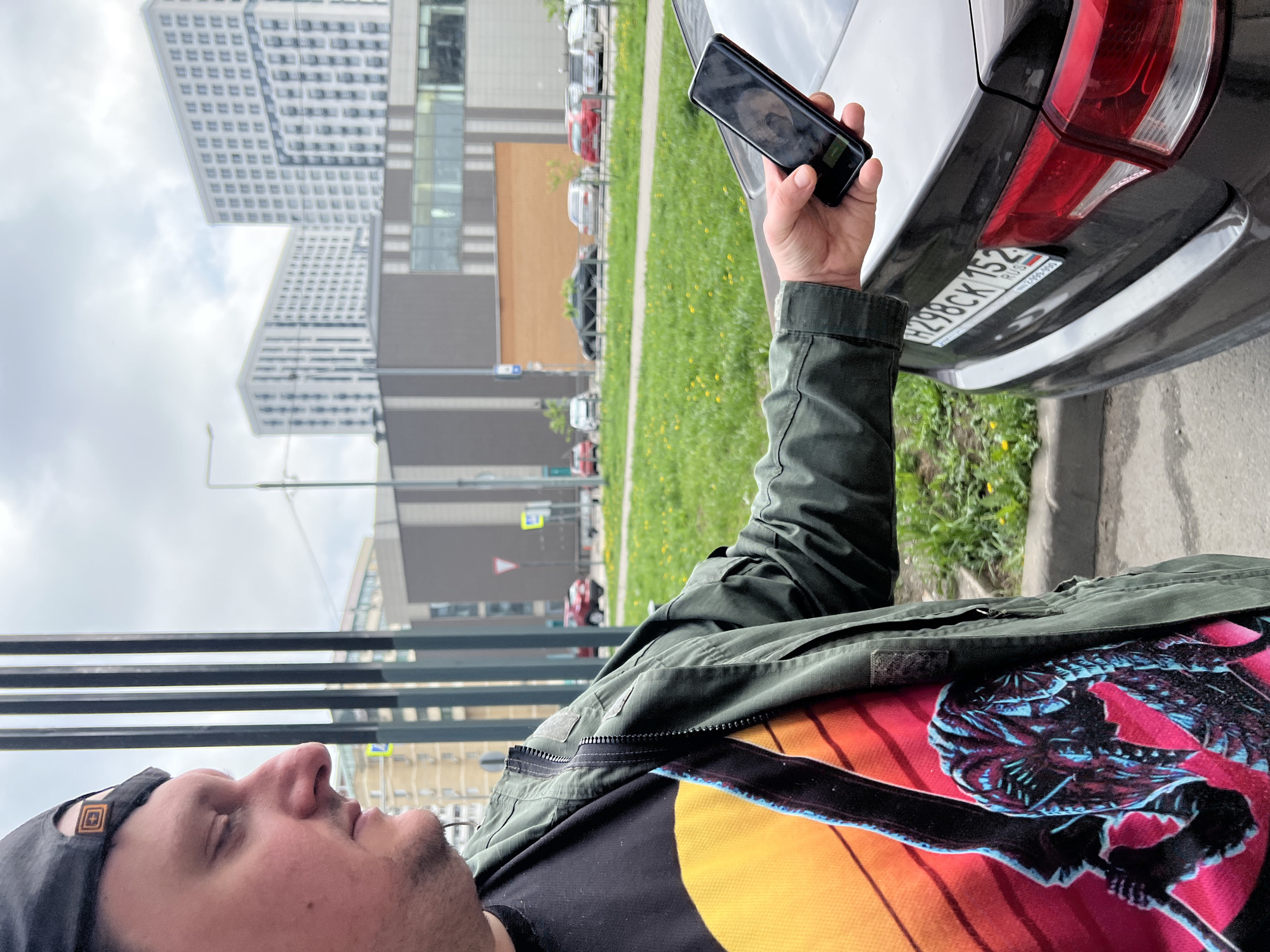 A man in a black cap and an olive green jacket is standing next to a grey car in Mytishchi, Russia. The car's license plate reads "H298CK 152 RUS". He is holding a black smartphone in his right hand and appears to be taking a photo or video of the car's license plate. The background includes a grassy area with dandelions, a street with parked cars, and several buildings, including a high-rise residential complex with a distinctive stepped design. A road sign is also visible. The man is wearing a t-shirt with a sunset graphic and a stylized skull or monster design.