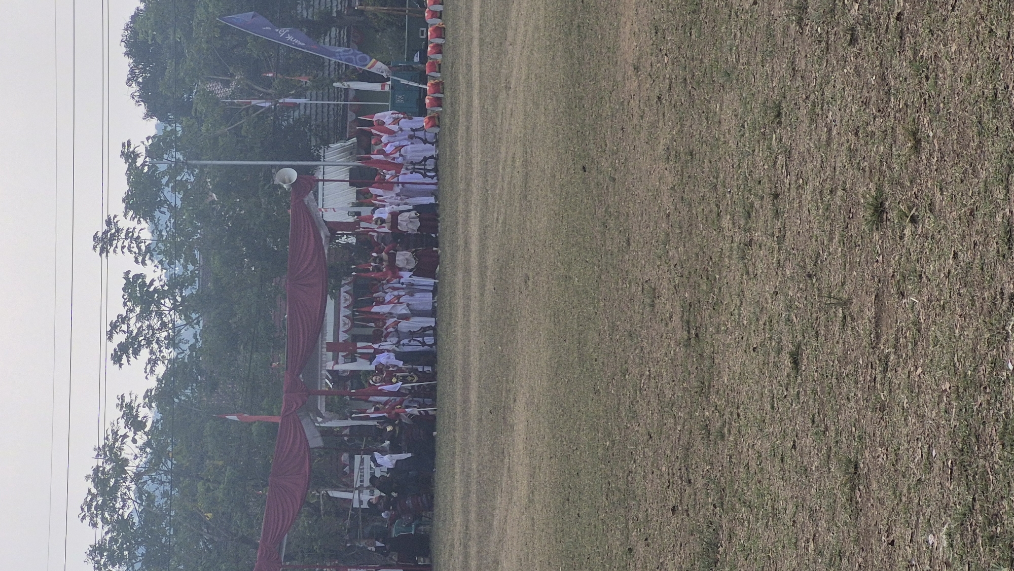 The image depicts an outdoor scene at a field, likely in Limbangan, Indonesia, where a group of people are gathered under a red canopy. The individuals are mostly dressed in white attire with red accents, many holding flags. The group appears to be part of a procession or ceremony, with some individuals wearing more elaborate traditional clothing. In the background, trees and a cloudy sky are visible. To the right of the canopy, several red objects are lined up on the ground, possibly part of the event setup. A flag with blue and white elements is also visible. The foreground is a dry, grassy field.