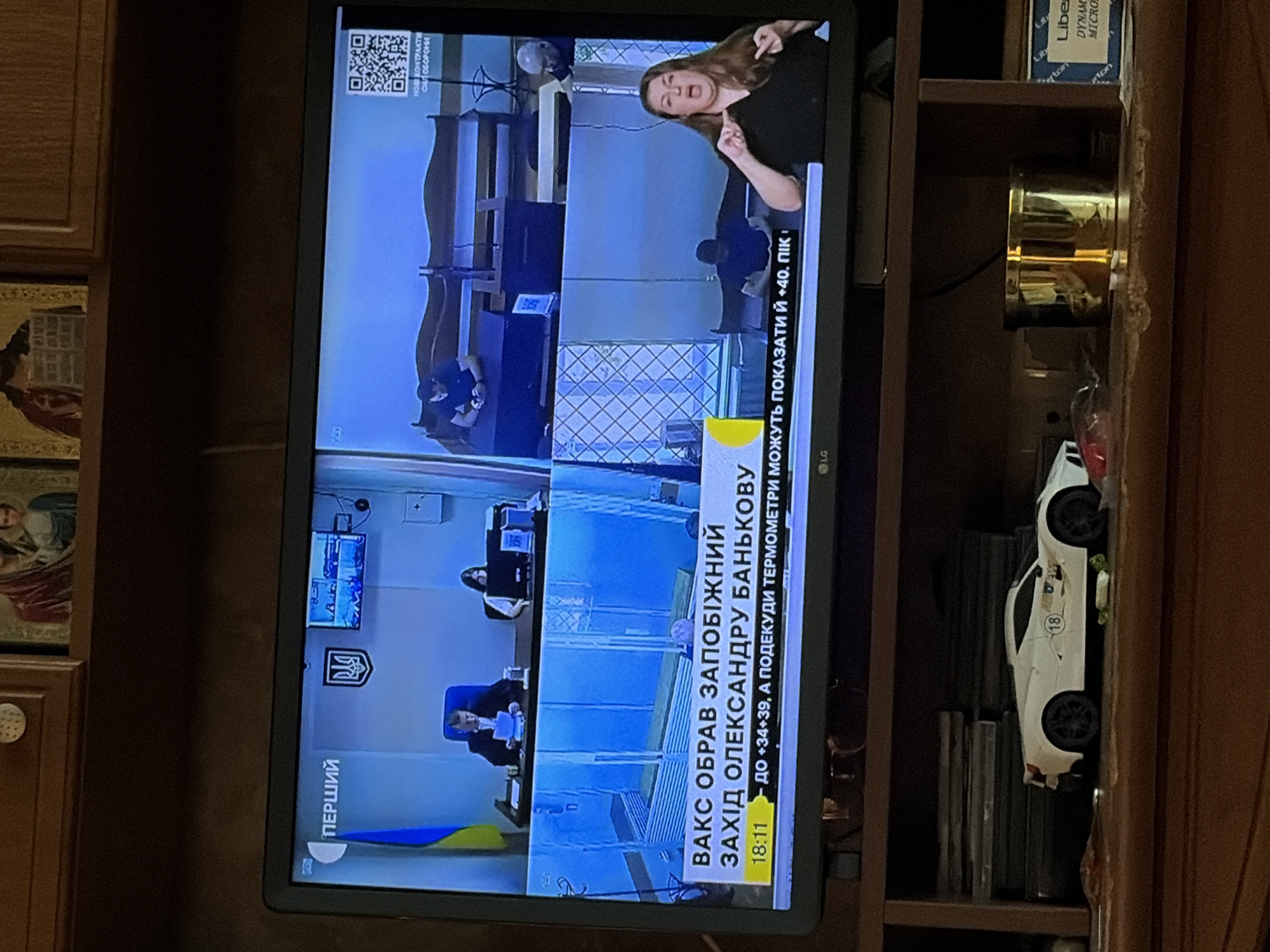 The image shows a television screen displaying a news broadcast. The broadcast is in Ukrainian, indicated by the text "ПЕРШИЙ" in the upper left corner. The screen is divided into multiple sections, showing a courtroom setting with people in suits, a woman signing, and a QR code. There is also a ticker tape at the bottom of the screen with Ukrainian text. The television is placed on a wooden shelf, below which is another shelf holding various items, including a white toy sports car, a golden container, and some books or DVDs. In the background, above the television, religious icons are visible. The overall scene appears to be a domestic setting, likely a living room.