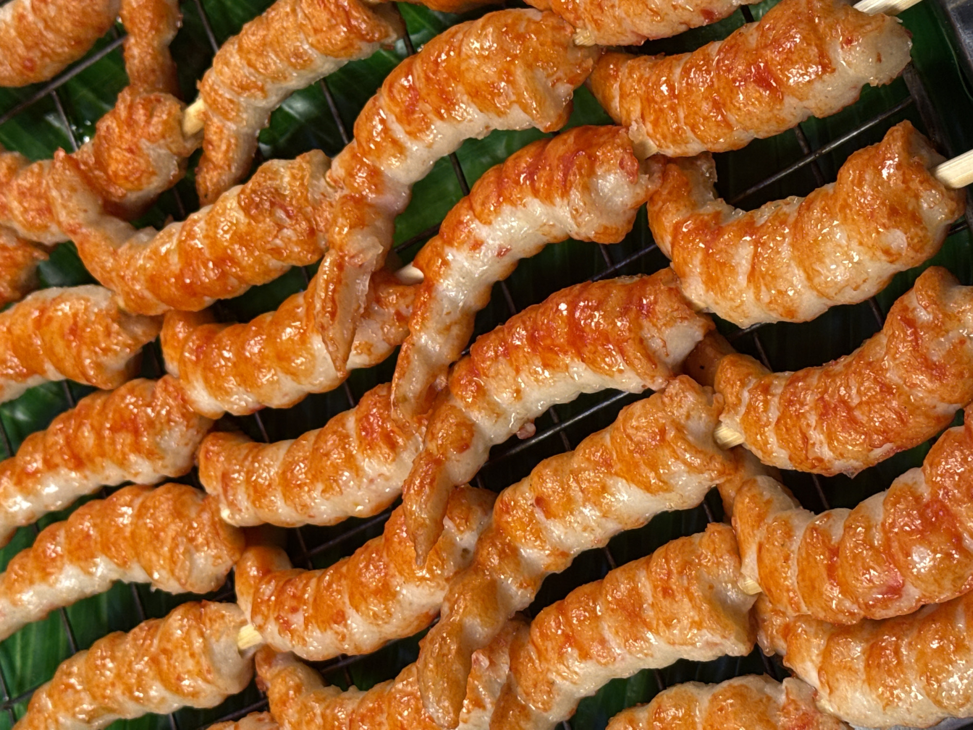 The image shows a close-up, top-down view of several skewers of grilled shrimp laid out on a dark green, textured surface, likely banana leaves, which are resting on a black metal grill. The shrimp are curled and appear to be spiral-cut, with a glossy, golden-brown exterior, indicating they have been grilled or fried. The skewers are made of thin, light-colored wood. The lighting is bright, highlighting the glistening texture of the shrimp. The setting is likely an outdoor food stall or market in Karon, Thailand, given the visual cues of tropical ingredients and grilling preparation often found in such locations. No people are visible in the image.