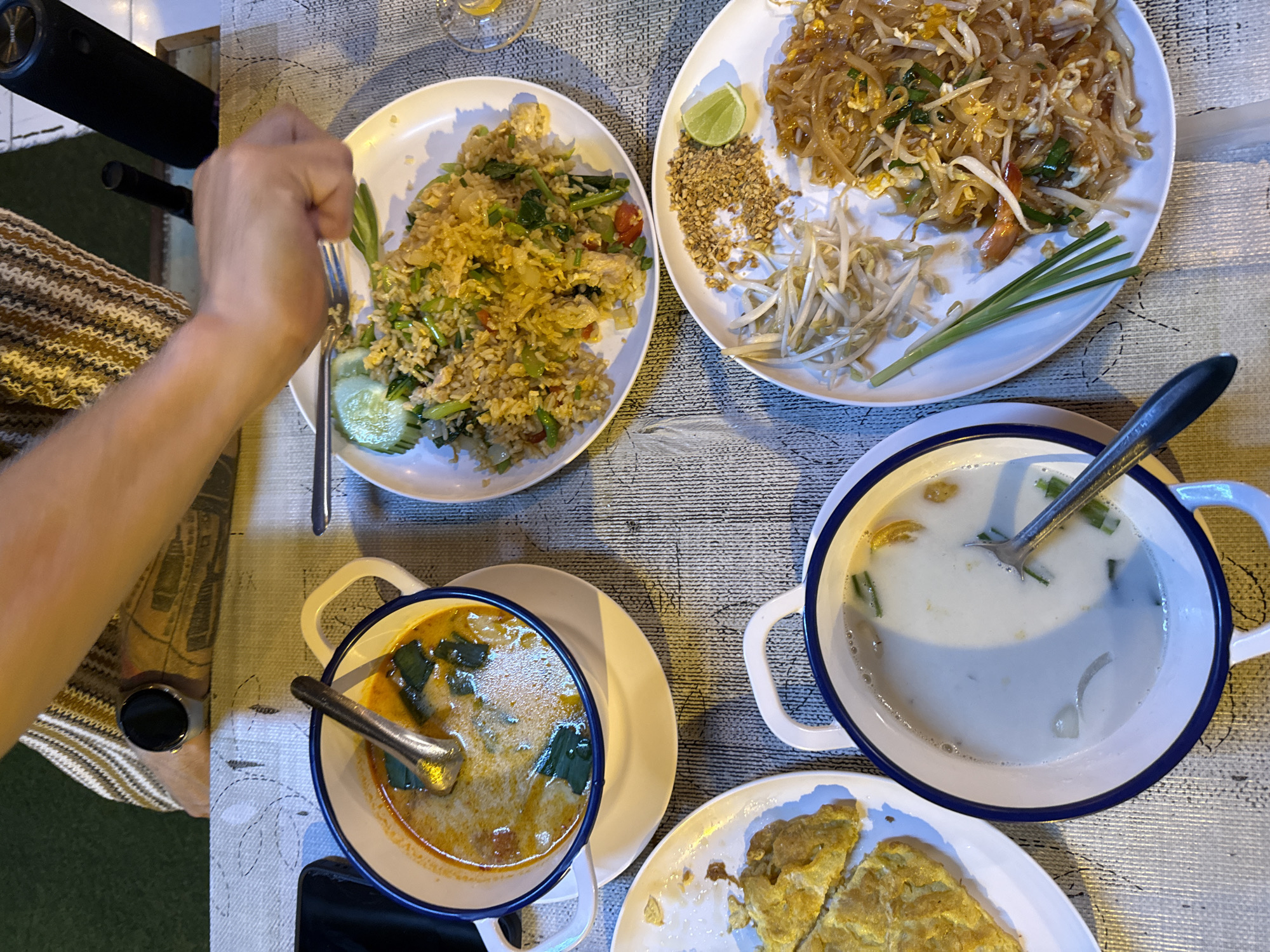 An overhead shot shows a table at an outdoor restaurant with several dishes of Thai food. A person's arm and hand are visible on the right side of the frame, reaching for a plate of fried rice. The table is covered with a textured, light-colored tablecloth. The dishes include a bowl of Tom Yum soup with lemongrass and chili, a plate of Pad Thai noodles with bean sprouts and crushed peanuts, a plate of fried rice with vegetables, a plate of what appears to be an omelet cut into pieces, and a bowl of creamy soup, possibly coconut-based. A fork is resting on the plate of fried rice, and a lime wedge and chopped peanuts are garnishing the Pad Thai. The lighting suggests it is evening, with shadows casting on the table. A black speaker is visible in the background, to the right of the person's arm.