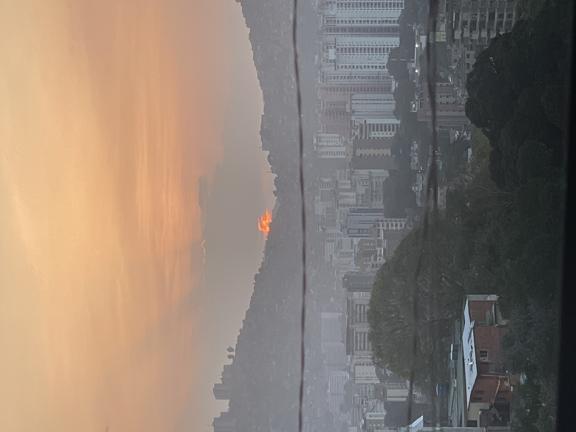 The image shows a cityscape in Caracas, Venezuela, viewed from an elevated position. The scene is dominated by high-rise buildings densely packed together, contrasted by tree-covered hills. In the distance, there's an orange glow possibly indicating fire activity on the mountain slopes. Powerlines are visible cutting across the view.