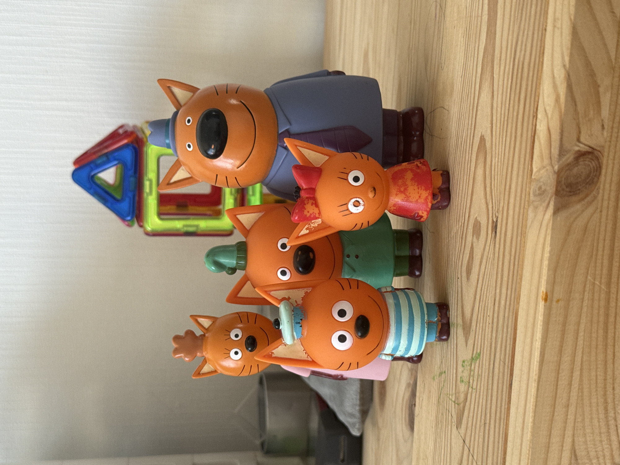 This image shows a collection of cat figurines arranged on a wooden surface. The figurines are brightly colored and appear to be from a children's cartoon. In the background, a set of magnetic building blocks is visible, forming a small house-like structure. The overall setting suggests a playful and indoor environment. There is no information in the image that allows for the determination of its location in Lyubertsy, Russia.