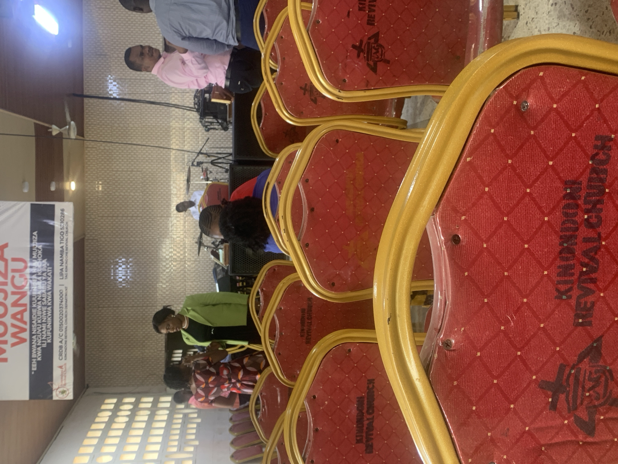 The image captures a scene within the Kinondoni Revival Church in Dar es-Salaam, Tanzania. Red chairs with gold trim fill the frame, each adorned with the church's name and logo. The chairs are arranged in rows, suggesting an auditorium or meeting hall setting. Attendees are visible, including women wearing dresses and jackets. In the background, a stage is set up with musical instruments, and a banner displays church details and a prayer in Swahili. Men wearing collared shirts are also visible in the background.