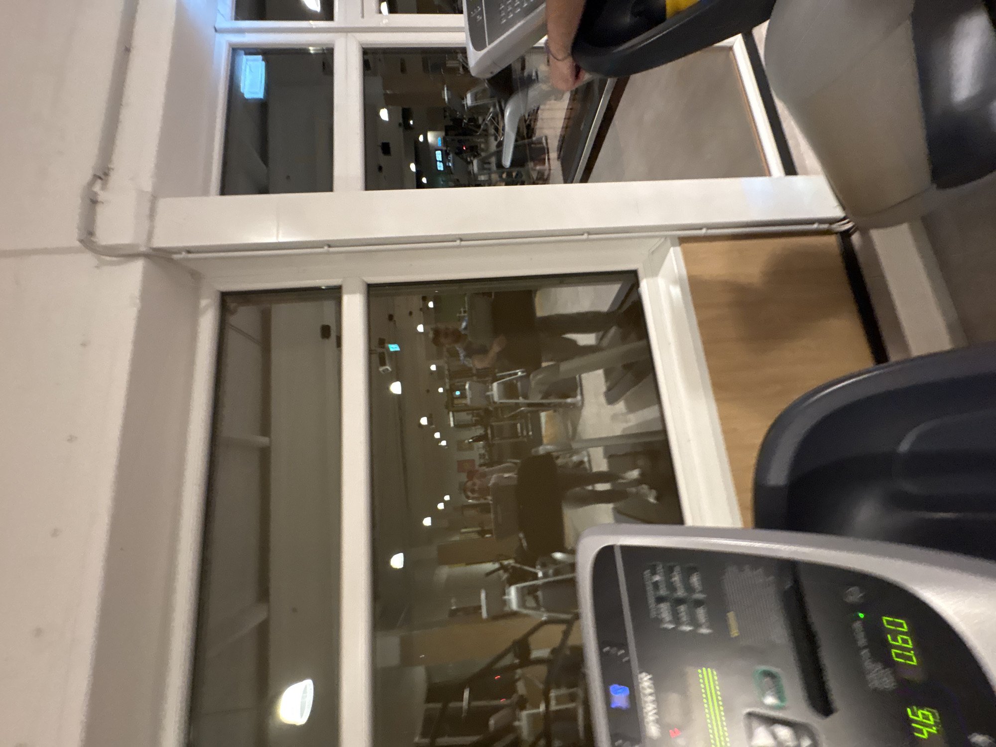 The image captures the interior of a gym in Mönchengladbach, Germany, as viewed through a window. Exercise equipment, including treadmills with illuminated consoles displaying metrics such as speed (4.6) and distance (0.60), dominates the foreground. Reflections on the window's surface show other gym members engaged in workouts. The environment is brightly lit and appears clean.