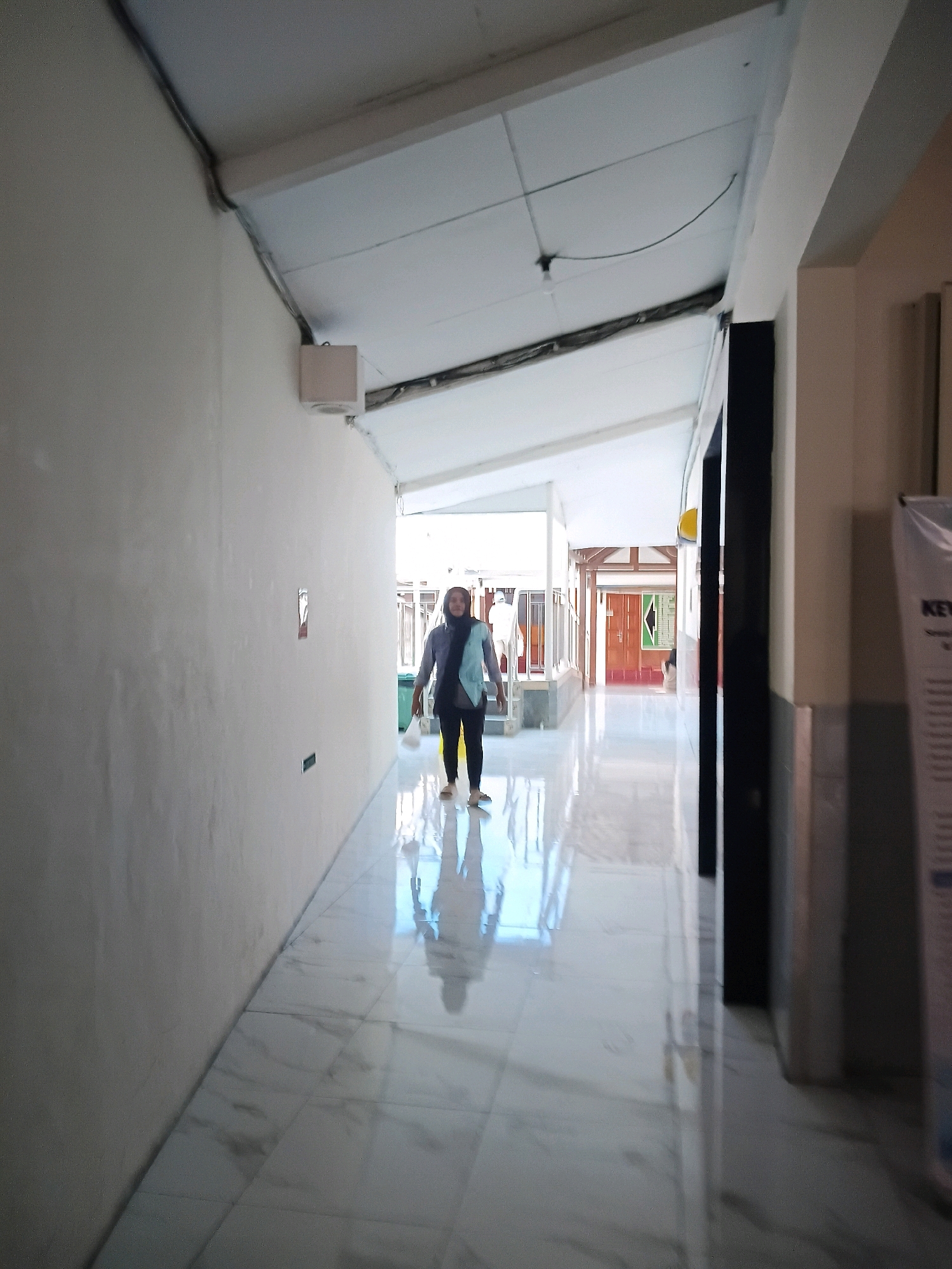 A young woman wearing a hijab and a light blue and grey outfit walks down a brightly lit, modern hallway. The hallway has a white ceiling and light grey walls. The floor is made of polished white tiles with a subtle marble pattern, reflecting the light and the woman's shadow. The woman is facing forward, her left hand holding a white object. In the background, there are doors and windows that lead to an outdoor area. On the right side of the hallway, a poster is partially visible. The scene appears to be in a public building, possibly an educational institution or a hospital, in Purwodadi, Indonesia.
