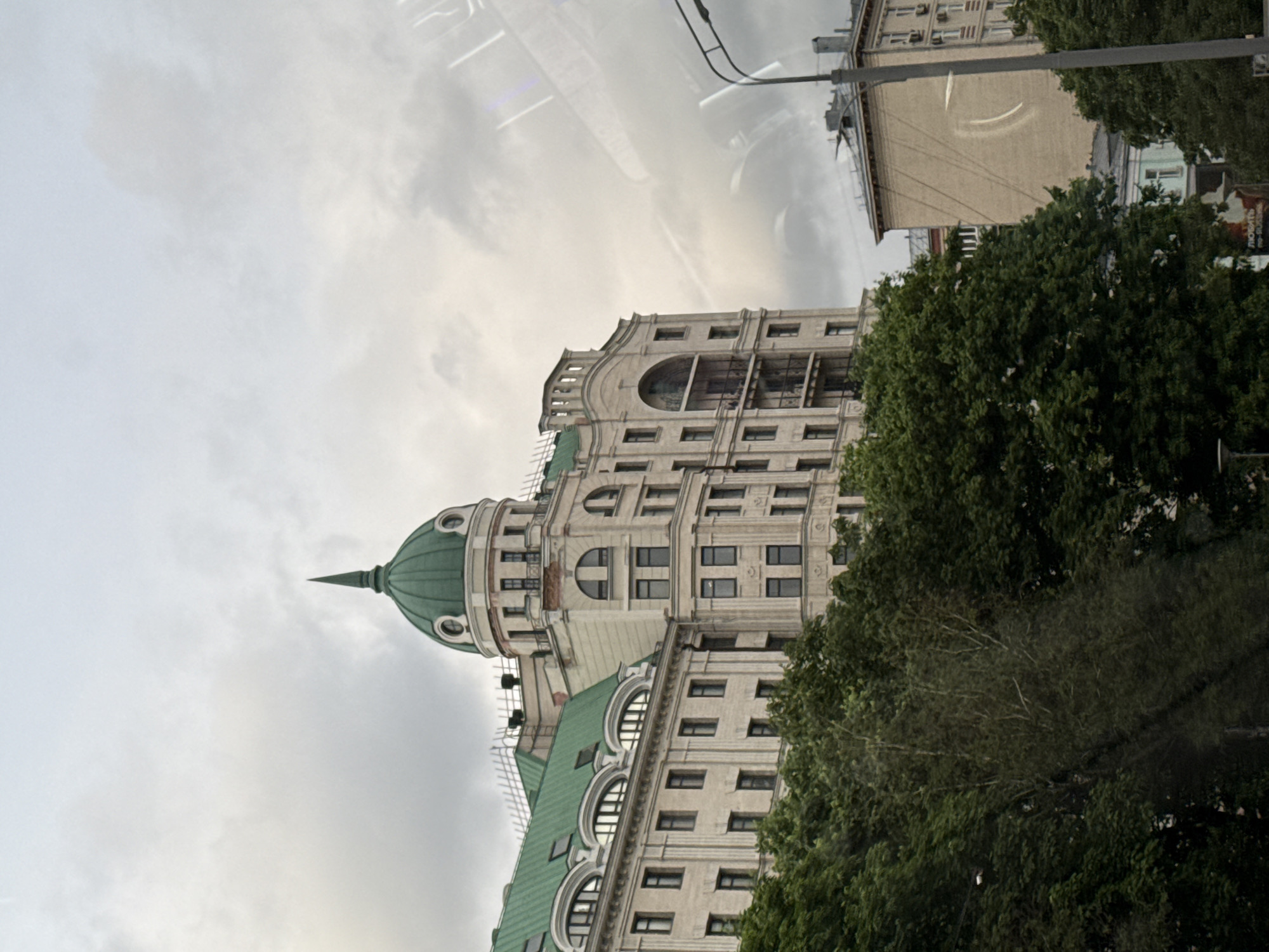 The image shows a tall building in Moscow, Russia. The building has a green roof and a distinctive dome. Trees are visible in the foreground, partially obscuring the building. A utility pole with wires is also visible near the top of the frame. The sky is overcast. Reflections are visible on the window that took this photo.