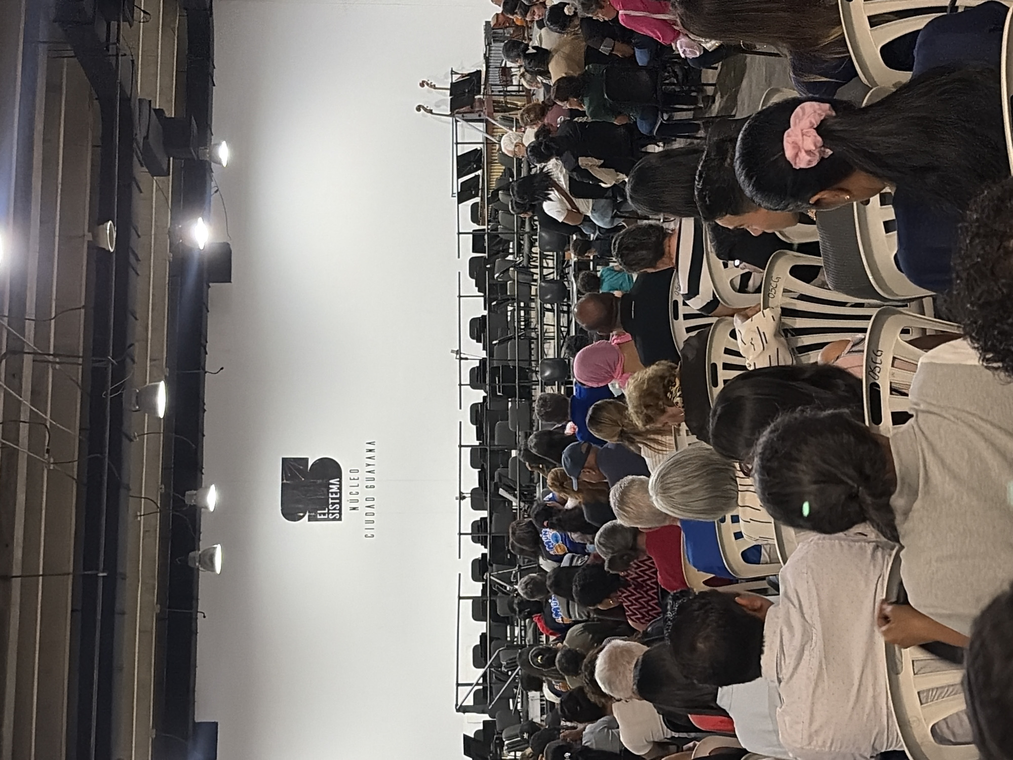 The image shows an audience seated in rows of chairs in what appears to be an auditorium or large hall. The wall in the background features the logo for "El Sistema, Nucleo Ciudad Guayana." The auditorium has lighting fixtures and visible structural elements on the left side of the frame. An orchestra is visible performing on a stage at the back of the hall. The audience members are looking towards the stage. The location is Ciudad Guayana, Venezuela, as per the logo.