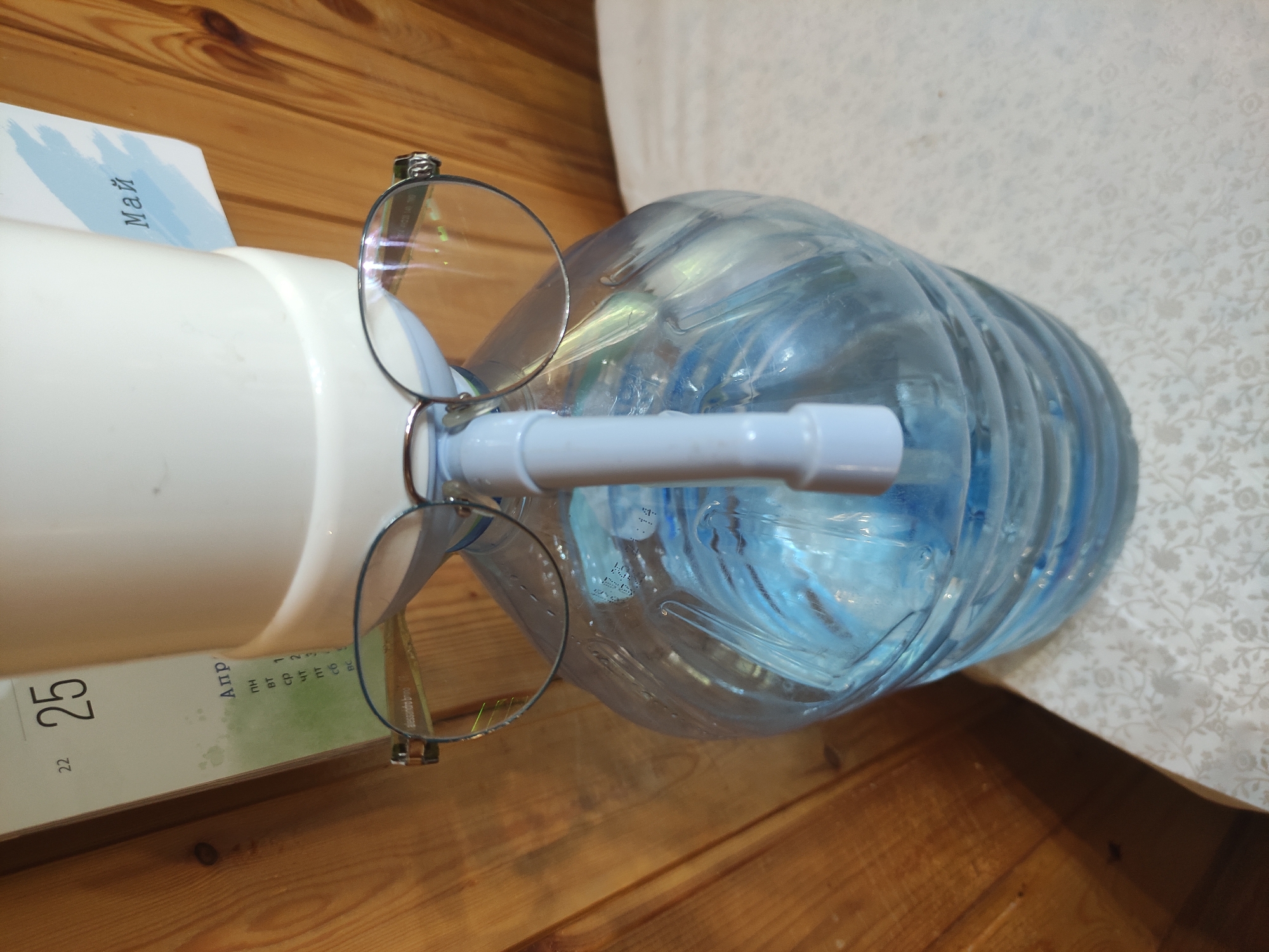 The scene depicts a large, transparent plastic water bottle affixed to a white water cooler. A pair of silver-framed glasses rests on the cooler's spigot. A calendar is positioned to the left, showing the date "25" for April and the month of May on a page behind it. The setting is indoors, on a wooden surface and a patterned tablecloth. No people are visible. The location is likely not in Kratovo, Russia, as the calendar has text in a different language than Russian.