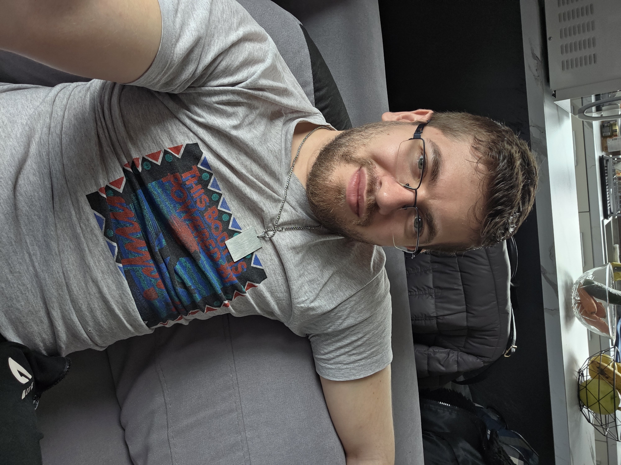 A man is lying on a gray sofa. He is wearing a gray T-shirt with a colorful geometric design on the front and a silver chain necklace. He has short brown hair, a beard, and glasses. Behind him is a black jacket hanging in a closet, and a fruit basket containing bananas and oranges is visible. The location is not identifiable as Yagodovo, Bulgaria.