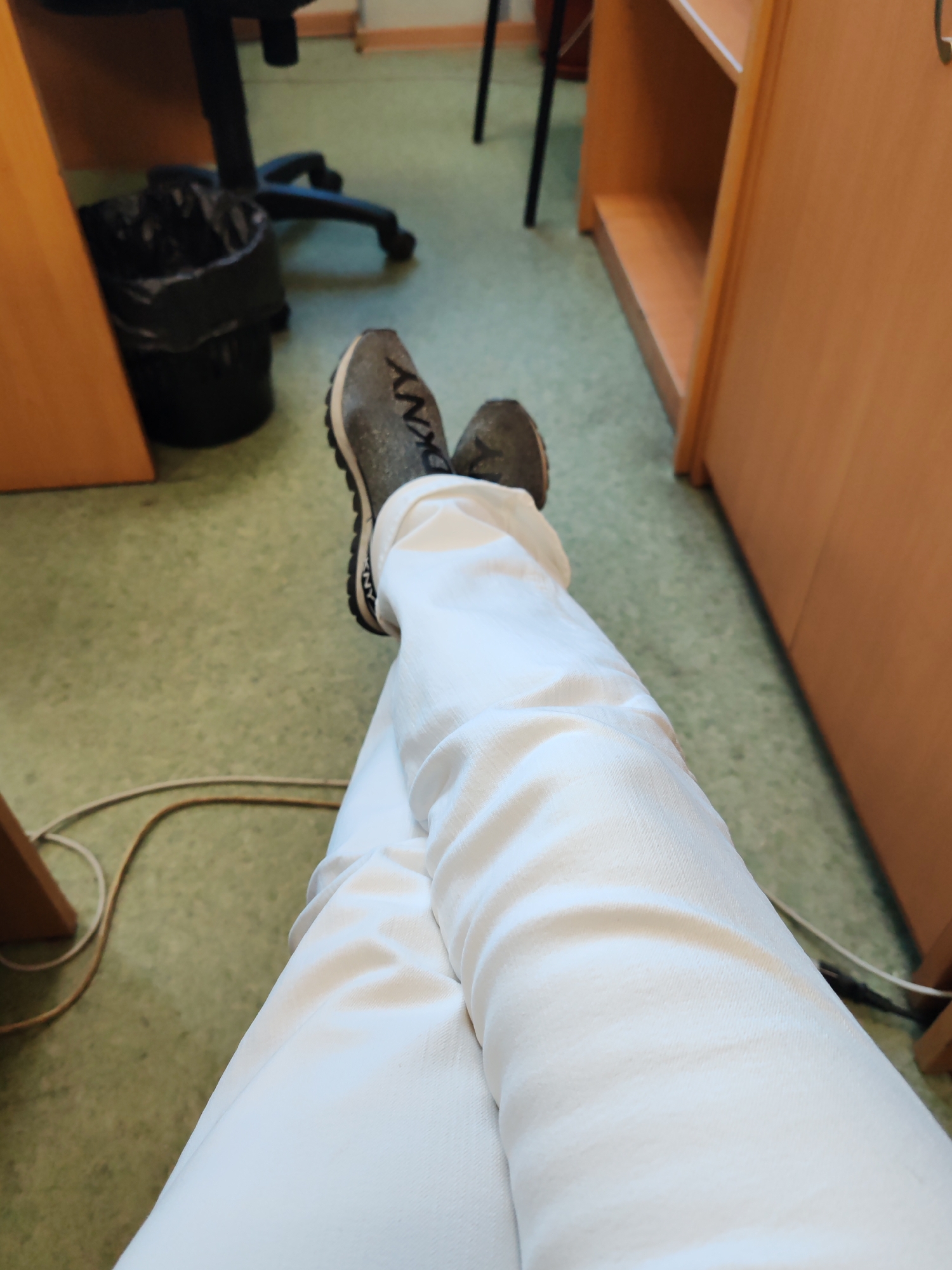 The image presents a first-person perspective, looking down at a person's lower legs and feet as they sit with their legs crossed. The individual is wearing clean white trousers or pants. On their feet are a pair of dark grey or green athletic shoes, with the brand name "DKNY" clearly visible in black letters on the side of the left shoe. The shoes have prominent black soles with textured treads.
The scene is an indoor environment, likely an office, classroom, or study area. The floor is a light green, speckled material, possibly linoleum or vinyl. Various objects are visible in the background, suggesting a functional setting. To the left, part of a black office chair base with wheels is seen next to a black trash can with a black plastic bag liner. On both the left and right sides of the frame, sections of light brown wooden furniture, appearing to be desks or shelving units, are present. A light-colored cable, possibly an electrical or network cord, trails across the green floor near the person's feet.
The image captures a moment of relaxation or casual posture, with the person seemingly taking a break while seated. No specific time of day or weather conditions can be determined from the indoor lighting. The visible text is the "DKNY" logo on the shoe.
