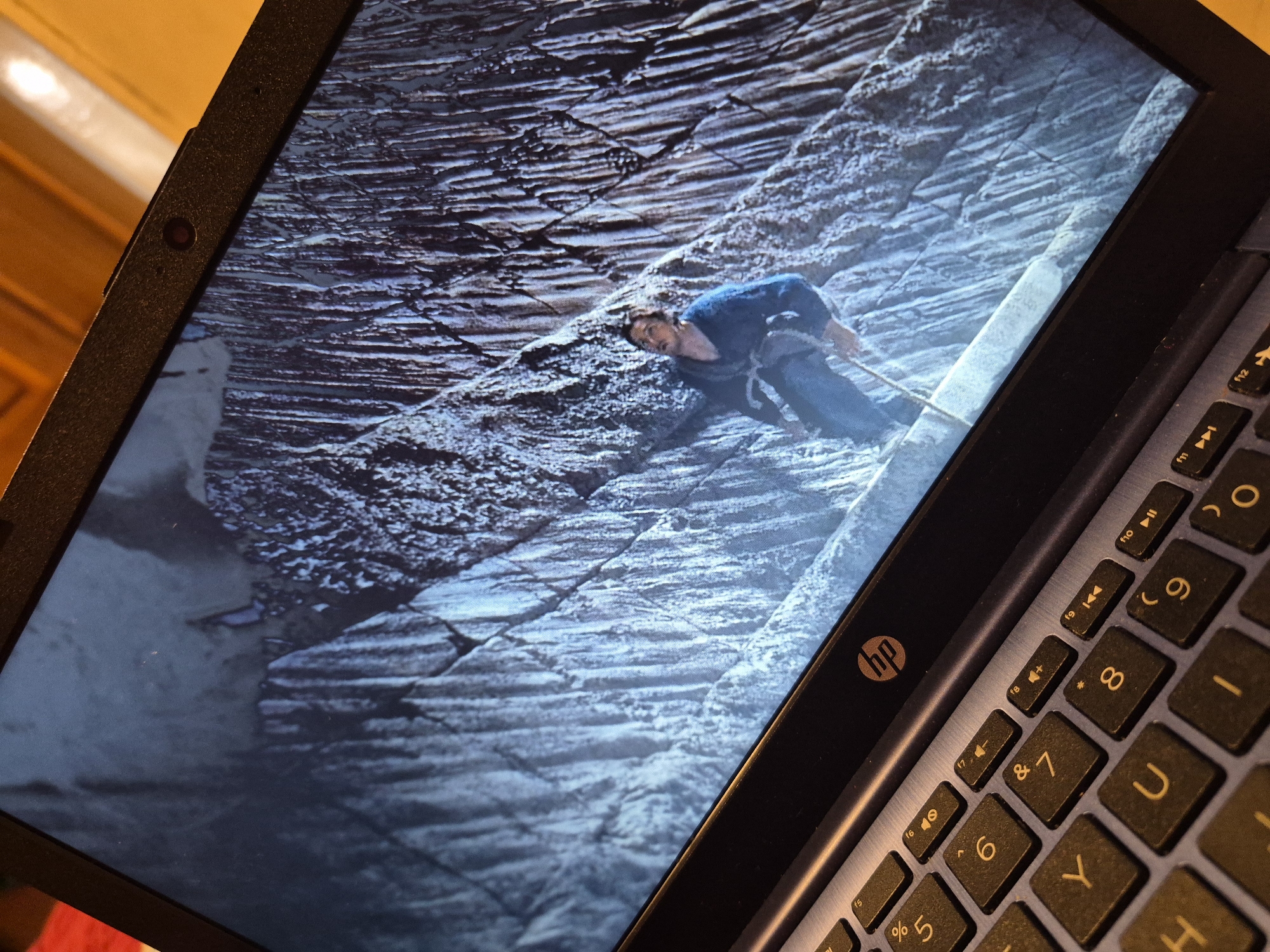 The image shows a laptop screen displaying a scene of a person rock climbing on a textured, grey, mountainous surface. The climber, wearing a blue outfit and a harness, is positioned on the left side of the screen, looking upwards. The laptop is open, with its keyboard visible in the foreground. The keyboard has an "HP" logo on the bezel above it. The lighting suggests an indoor setting, with warm light reflecting off the laptop's frame. No specific location in Sandwell, United Kingdom, can be identified from the image.