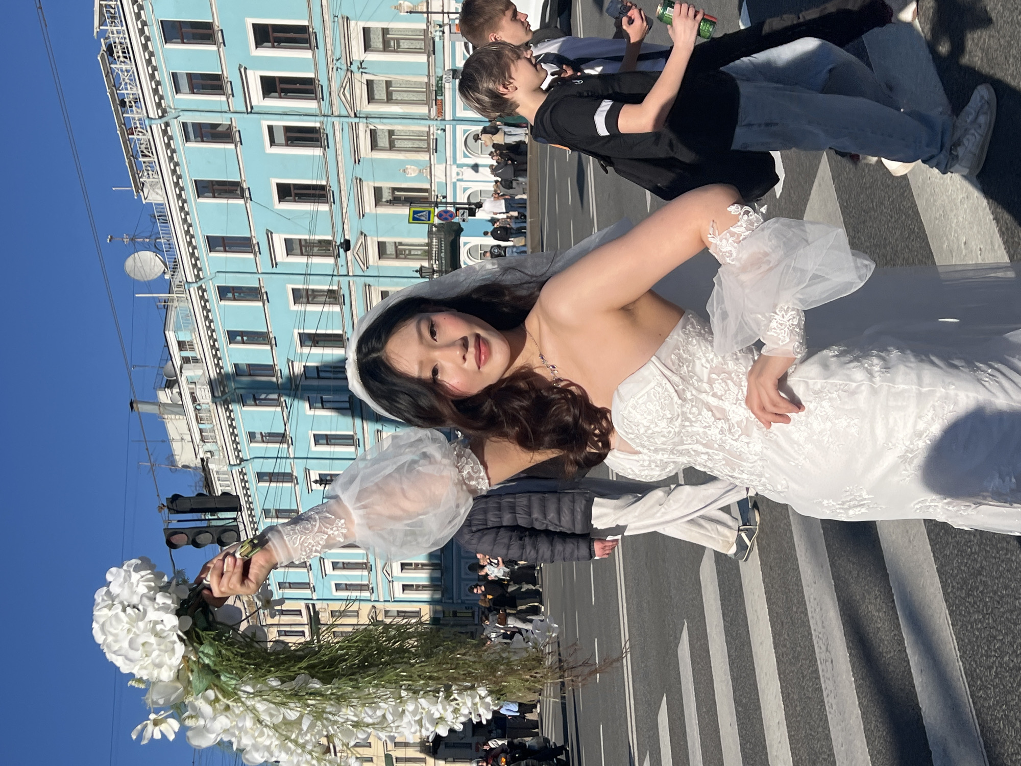 The image shows a woman in a white wedding dress holding a bouquet of white flowers, standing on a zebra crossing in the street. The buildings behind her are light blue with white window frames, characteristic of Saint Petersburg architecture. There are pedestrians on the street in the background. Two young men are passing behind her.