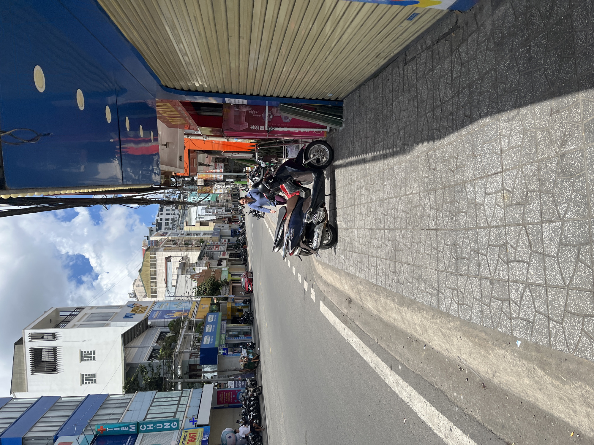 A street scene in Da Lat, Vietnam, features a wide asphalt road with white lane markings and a textured gray concrete sidewalk. Several motorcycles are parked along the sidewalk. On the right side of the image, a blue building with a metallic rolling security gate is prominent. A woman in a blue long-sleeved shirt and dark pants is walking along the sidewalk. Buildings with various architectural styles and signage line the street in the background, under a bright, cloudy sky.