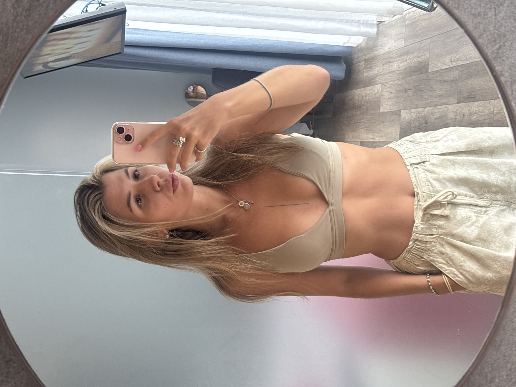 In a softly lit room with a pastel-colored wall and a wooden floor, a young woman with blonde hair tied back takes a selfie in a round mirror. She is wearing a beige bralette and patterned cream-colored shorts. Her right hand holds a pink smartphone, and her left hand is in her pocket. She is adorned with jewelry, including earrings, a necklace, several rings, and bracelets. The background shows a window with blue curtains and a television screen displaying an image. The overall impression is one of casual domesticity.