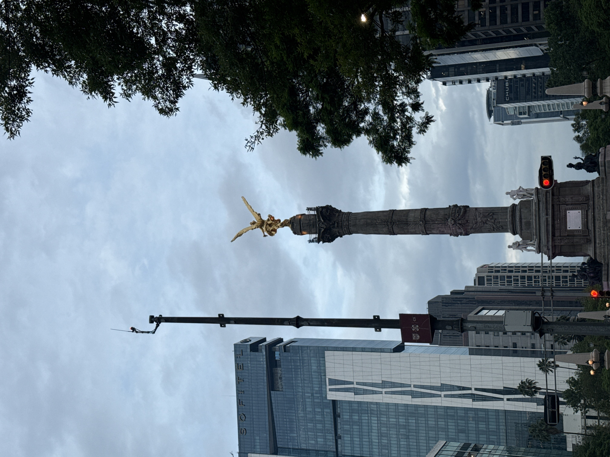 The image shows the Ángel de la Independencia monument in Mexico City, Mexico. The monument is a tall column topped with a golden statue of a winged Victory figure. Below the statue, the column is decorated with carvings and sculptures. Buildings are behind the monument. A stoplight is red. The sky is cloudy. A tree branch is at the top of the image.
