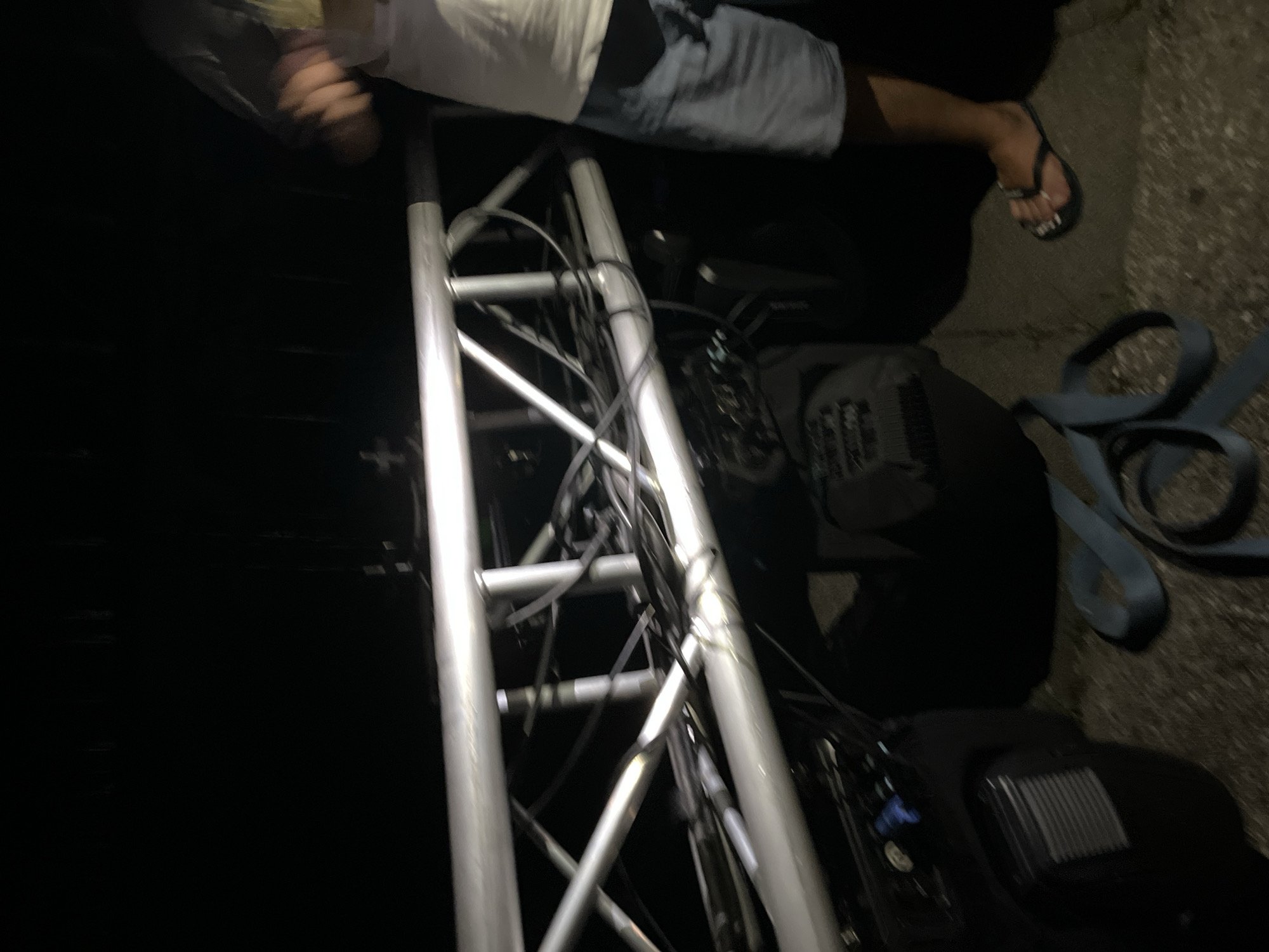 A low-angle, blurry photograph taken at night depicts a section of a silver aluminum truss system. Cables are bundled and attached to the truss. Below the truss, partially visible black equipment cases are present. A person is standing to the right of the truss, wearing a white t-shirt and blue shorts, and flip-flops. The ground appears to be paved. A blue strap lies on the ground in the foreground. The image is dark, with artificial lighting creating highlights on the truss.