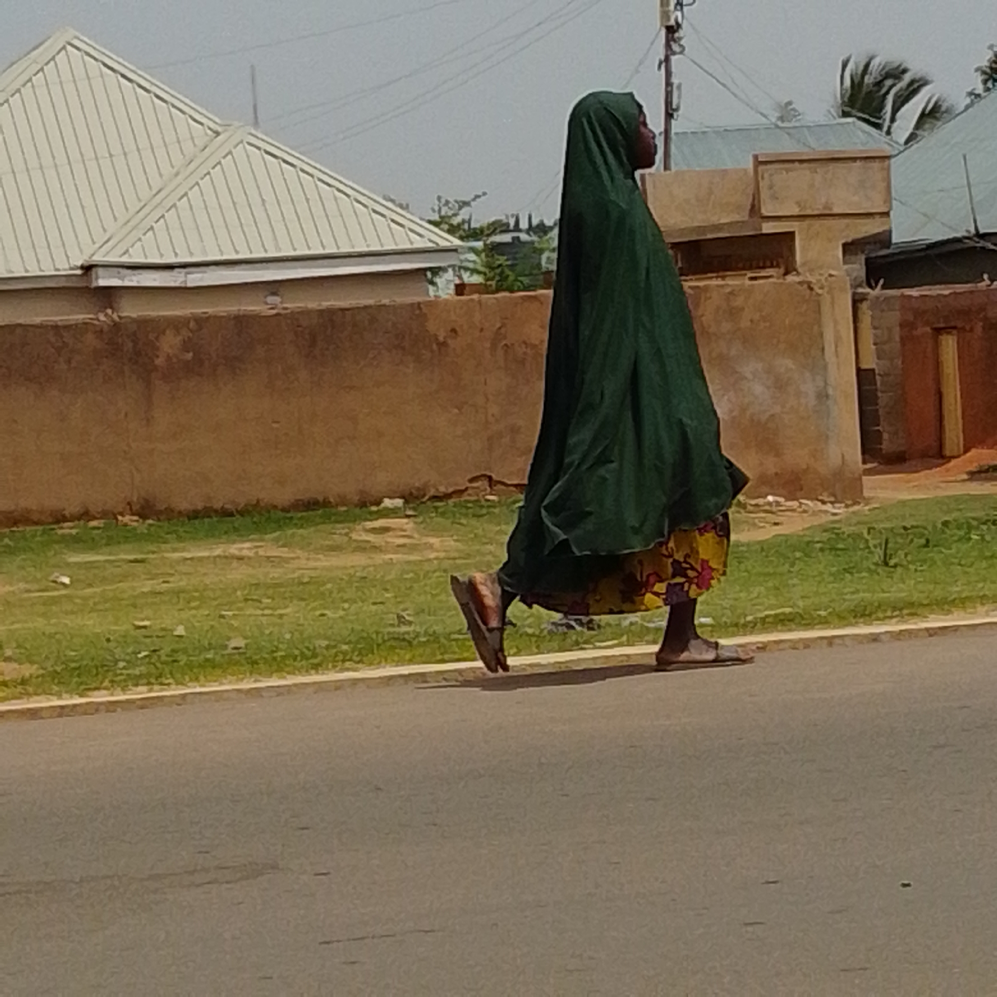The image depicts a person walking on a paved road adjacent to a grassy area. The person is wearing a dark green hijab and a patterned skirt. They are wearing sandals on their feet. In the background, there are buildings with light-colored roofs, and a brown wall. The sky is overcast. The scene appears to be in Gombe, Nigeria, based on the architectural style and vegetation.