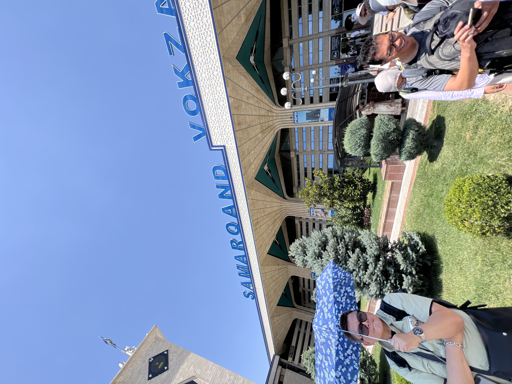 The image captures the Samarkand Railway Station in Samarkand City, Uzbekistan, on a sunny day. In the foreground, a man wearing sunglasses and a light-colored short-sleeved shirt is holding a blue umbrella adorned with white floral patterns. He has a backpack and is wearing a watch and a bracelet. To his right, a trimmed evergreen tree stands on a patch of grass. In the background, the grand facade of the train station is visible, with prominent blue lettering spelling out "SAMARQAND VOKZAL". The architecture features arched entrances and ornate detailing. Several other people are seen in the vicinity of the station entrance, some with backpacks and hats, suggesting they are travelers. The sky above is clear and blue.