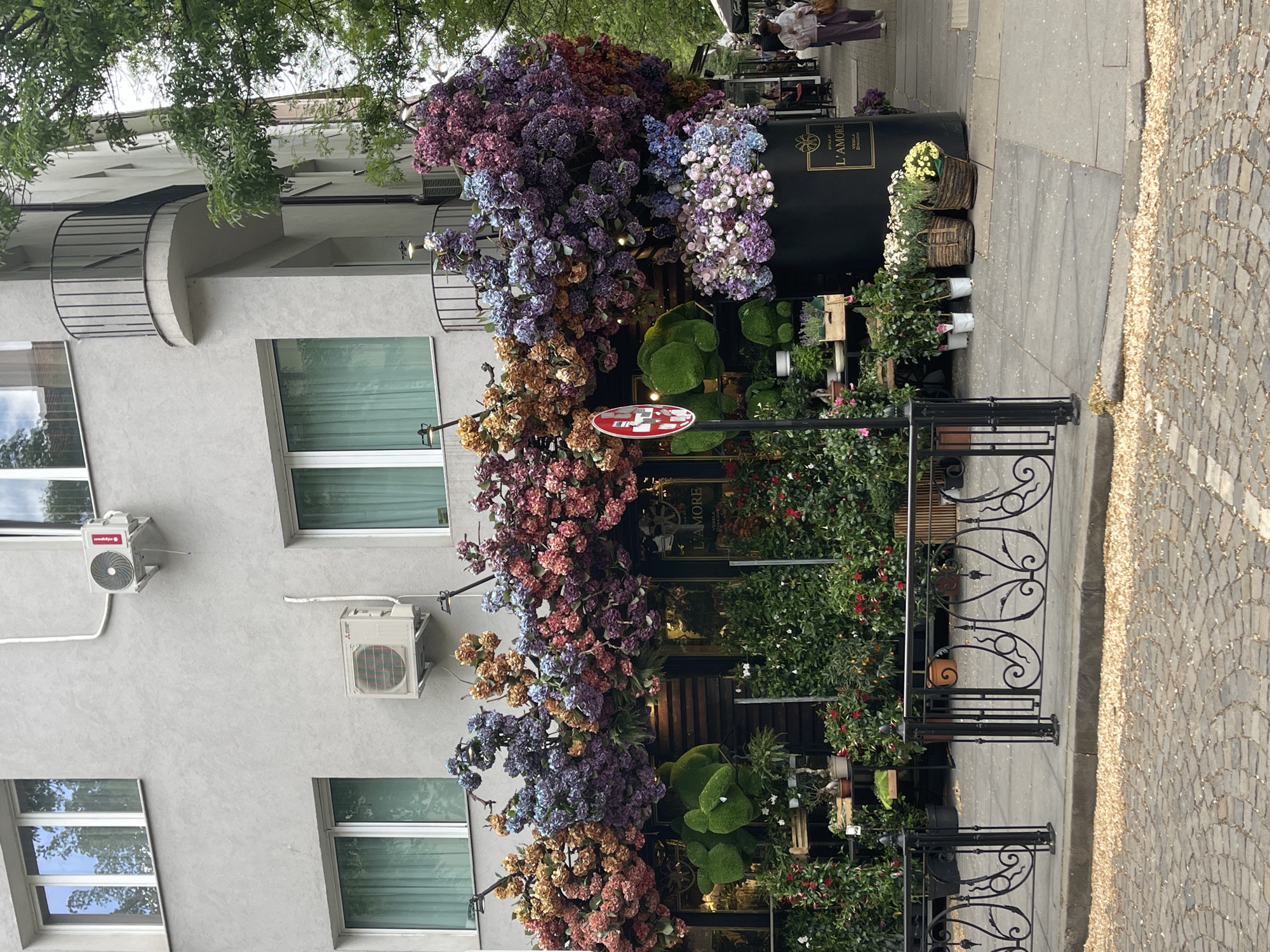 The image depicts the exterior of "L'AMORE," a florist shop in Sofia, Bulgaria. The shopfront is heavily decorated with flowers of various colors, including purple, blue, pink, and orange. Green foliage and topiaries are also visible. A wrought-iron fence separates the shop's display from the sidewalk.
The shop is situated in a gray building with multiple windows, and two air conditioning units are mounted on the facade. The sidewalk is paved with cobblestones. Two pedestrians are partially visible in the background.