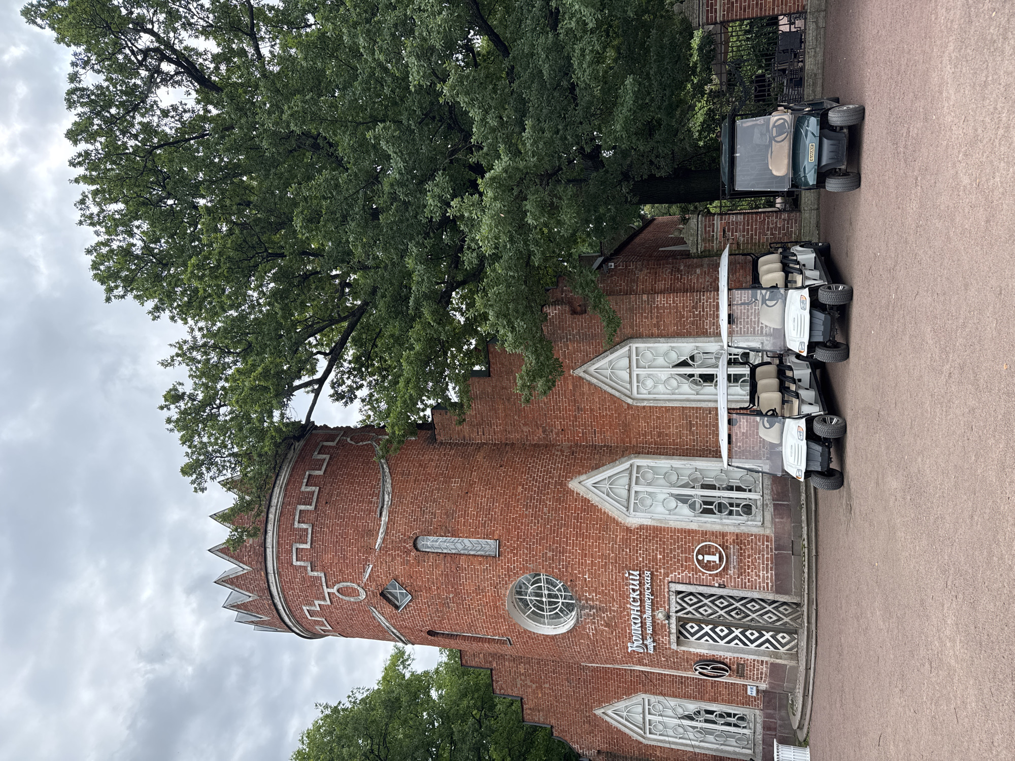 The image shows the "Volkonsky" cafe-confectionery building in the city of Pushkin, Russia, a red brick structure with a turret topped with a decorative crown-like element. The building features Gothic-style windows and a round window with an intricate design above the entrance. In front of the building, there are three golf carts parked on a reddish gravel surface. Two white golf carts are parked side-by-side, and a dark green golf cart is parked to their right. A large, leafy tree extends from behind the building, its branches overhanging the structure. The sky is overcast with clouds.