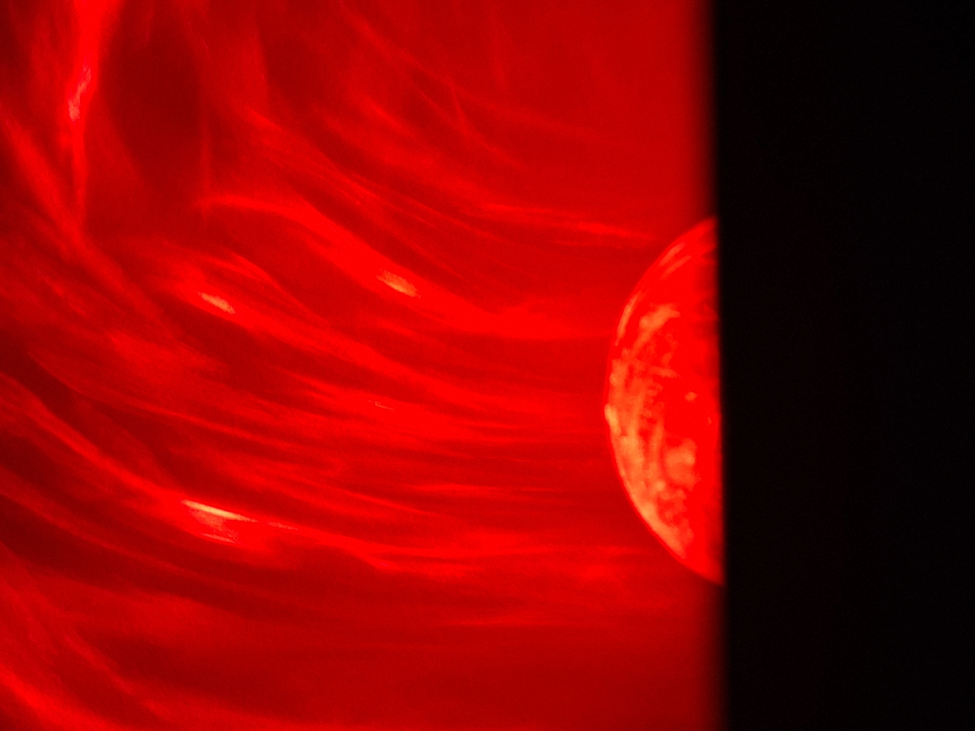The image shows a red and black abstract composition. The left side of the image is a vibrant red with swirling, streaky patterns. Within this red area, there is a vaguely spherical shape with lighter, orange-toned patches visible on its surface. To the right, a stark black area occupies a significant portion of the image, creating a sharp contrast. No people, recognizable objects, or identifiable environmental details are present to connect the image to Novi Sad, Serbia.