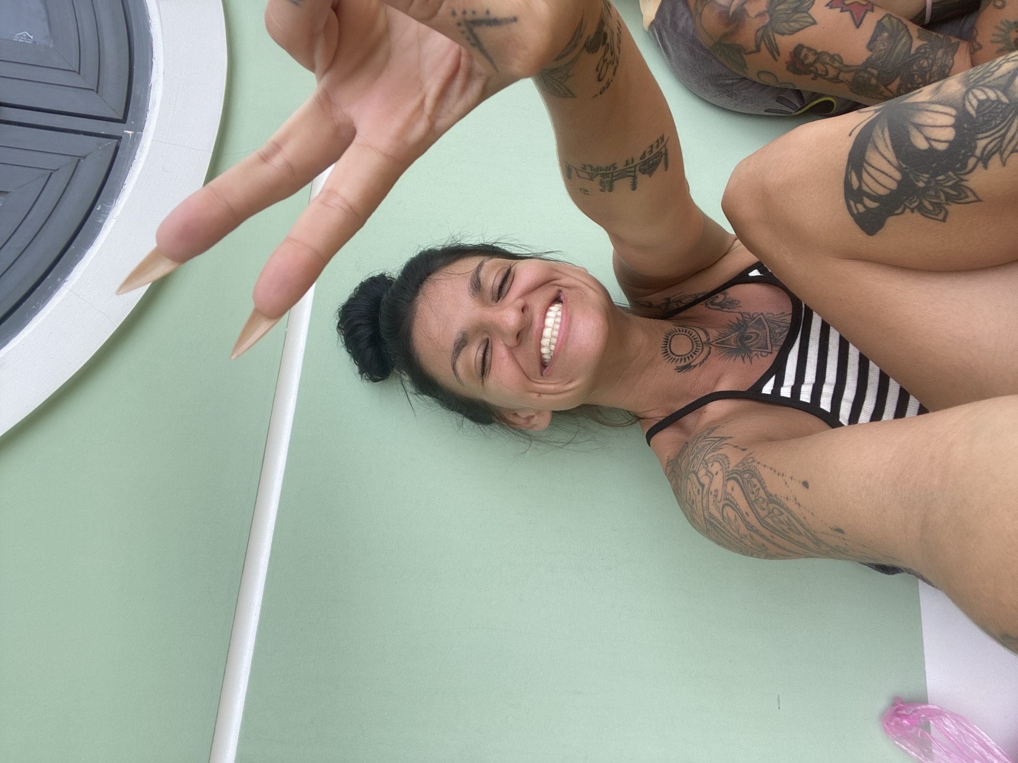 The image features a woman with dark hair, styled in a bun, lying on a light green surface. She is smiling broadly with her eyes closed, her head tilted back. Her body is adorned with numerous tattoos, including a butterfly on her arm and intricate designs on her chest and other limbs. She is wearing a striped bikini top. Her right hand is raised, with two fingers extended in a peace sign, and her fingernails are long and light-colored. The background shows a white rounded structure on the left, possibly part of a boat or a building. The overall mood is cheerful and relaxed. The scene is well-lit, suggesting it's daytime, possibly outdoors, given the location context of Mui Ne, Vietnam. A pink plastic bag is visible in the lower right corner. Visible text includes "KEEP IT SIMPLE" tattooed on her arm.
