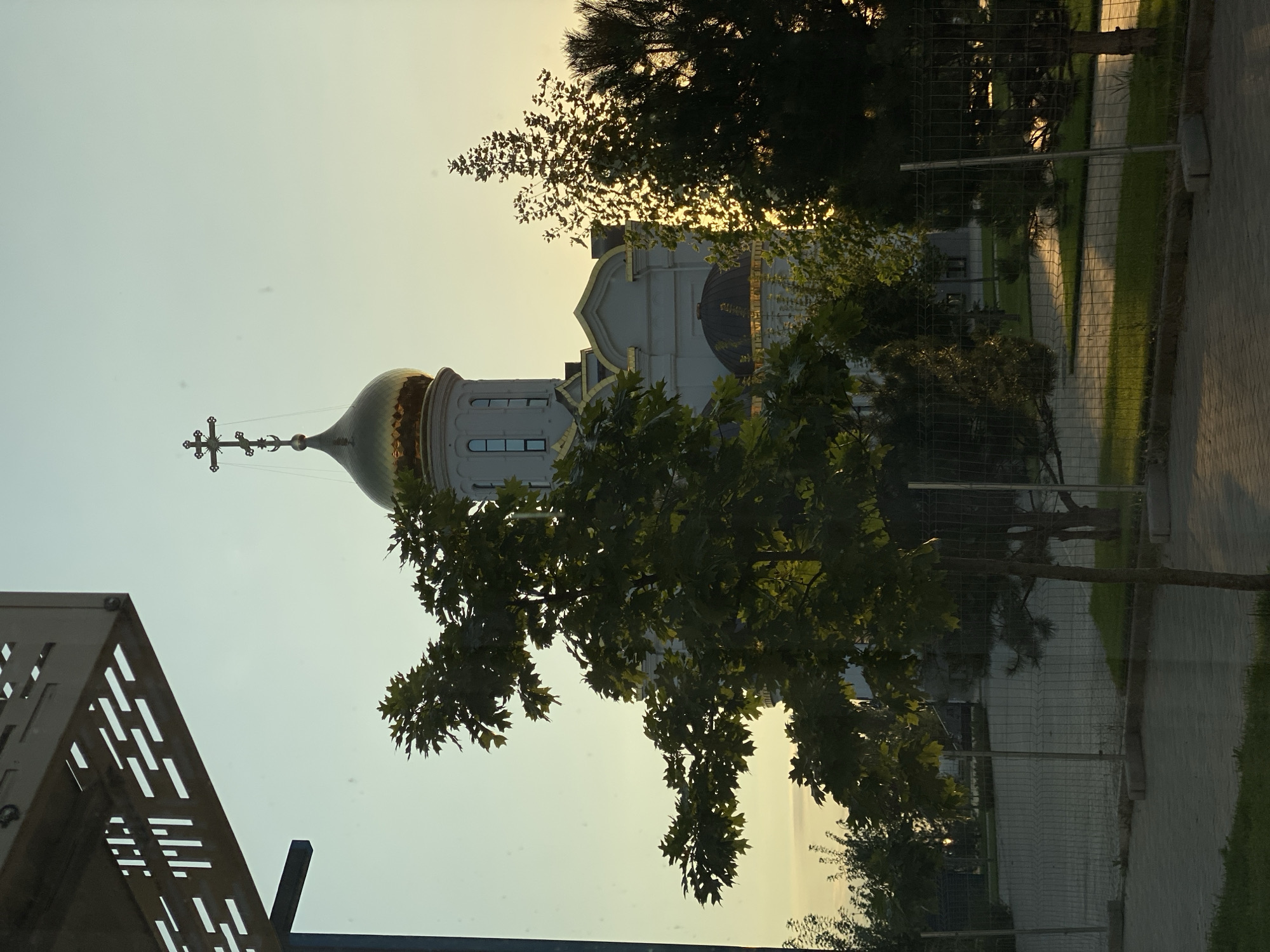 The image captures a view of the Church of the Ascension in Agronom, Russia, with a golden dome and cross prominently displayed against a pale sky. In the foreground, a tree with dark green leaves partially obscures the church. To the right, a dense coniferous tree is illuminated by the sun, creating a lens flare effect. A modern metal fence runs across the middle ground, separating a paved area from a grassy lawn. The overall lighting suggests it is either sunrise or sunset, casting long shadows and a warm glow on the scene.
