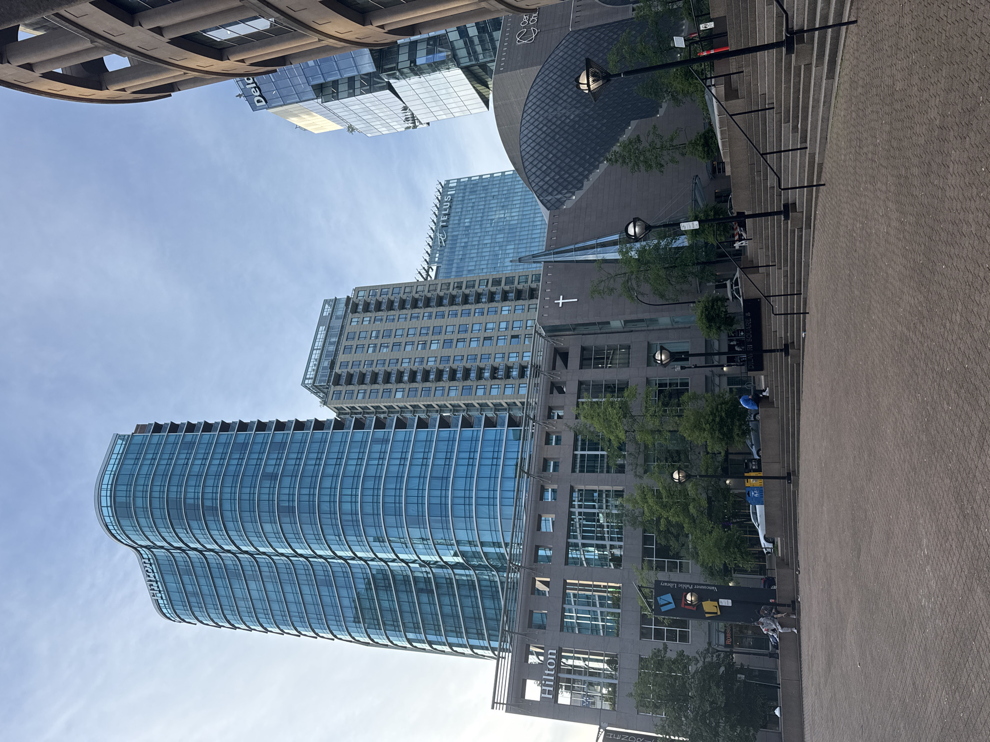 The image shows a view of downtown Vancouver, Canada, captured from street level. The scene includes several buildings, including a modern glass-clad hotel tower with "Hilton" signage, and other skyscrapers. A building with "Deloitte" signage is visible in the background.
There is a building with a cross, and a structure that may be a church or other similar gathering place.
In the lower right, there is an area paved with cobblestones, appearing to be part of a plaza. There are steps and lampposts lining a stairway. A few people are walking near a building with a "Vancouver Public Library" sign.