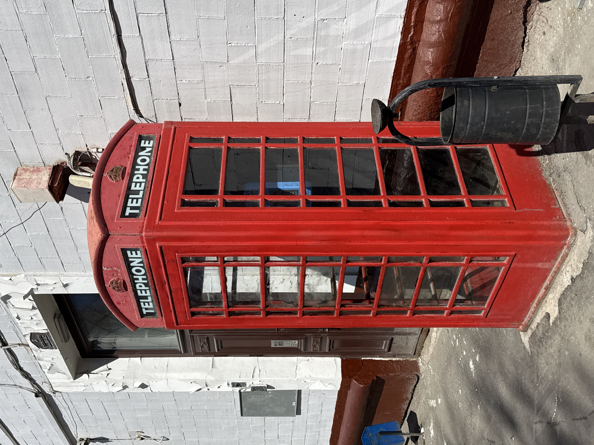 A red telephone box stands in Khimki, Russia. It has a black garbage can attached to its side. The telephone box is positioned in front of a white brick wall with peeling paint. A dark wooden door and a grey utility box are visible behind the phone booth. The scene is illuminated by sunlight.