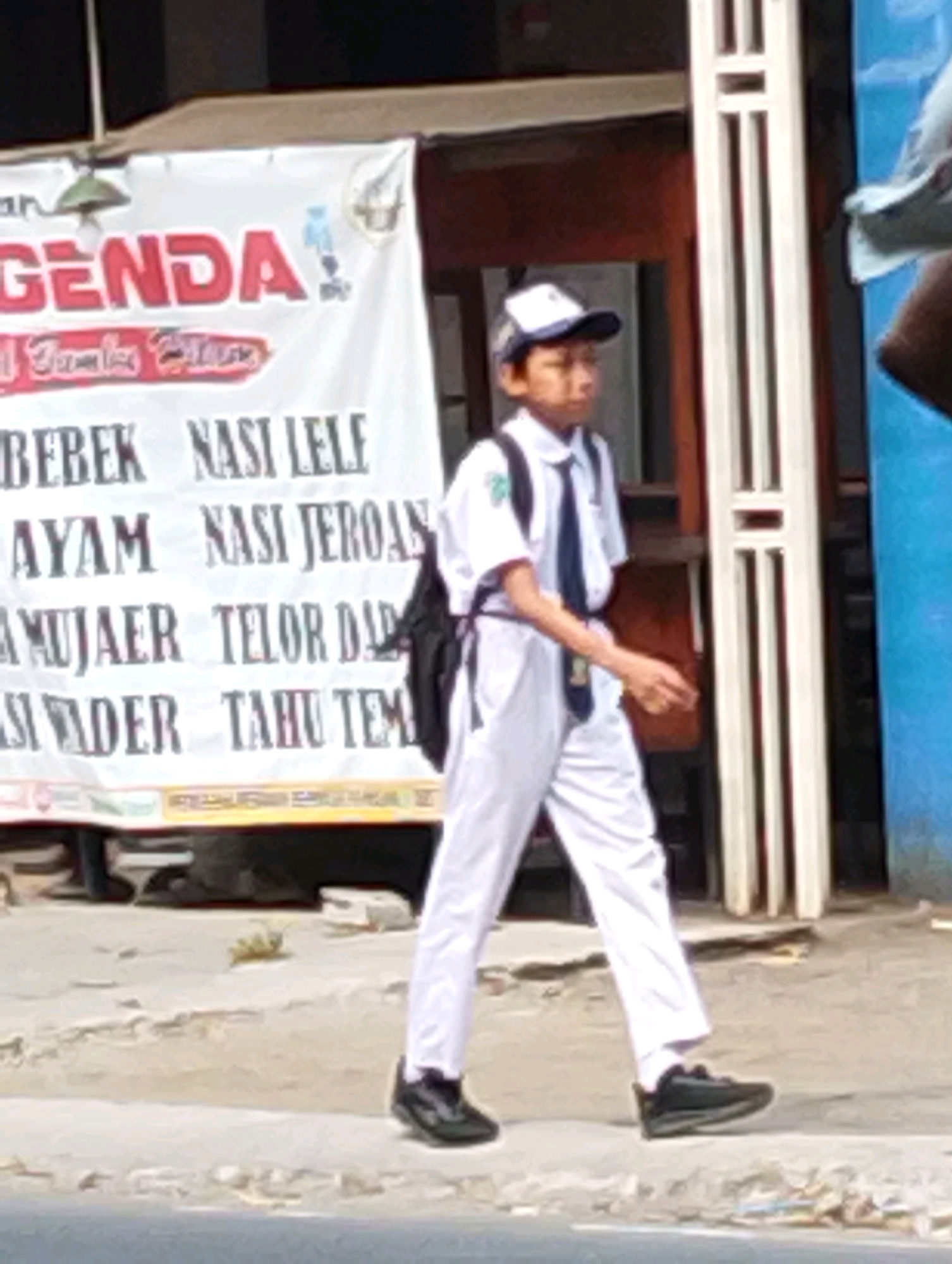 A young boy wearing a white school uniform, a blue tie, and a white cap walks on a sidewalk in front of a banner. The banner displays red text that reads "AGENDA" at the top and several lines of smaller black text below, including "BEBEK NASI LELE" and "AYAM NASI JEROAN". The boy carries a black backpack. To the right of the boy is a white structure with vertical openings. The background shows a building and a blue wall. The ground is paved and appears worn. This scene is located in Manaruwi, Indonesia.