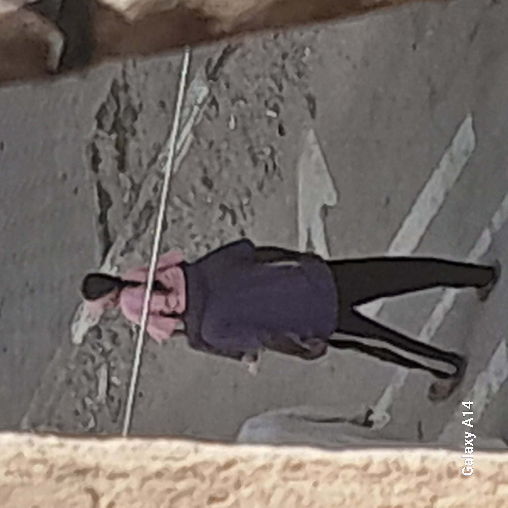 A person is walking away from the camera, carrying a child on their shoulders. The child is wearing a pink shirt and has a braid. The adult is wearing a dark shirt and black pants with white stripes. They are walking on a paved surface that appears to be a street, with some debris and a white line painted on it. To the left of the frame, a textured wall or structure is visible. The background is a blurred grey and brown textured area, possibly rough terrain or buildings. The image is marked with "Galaxy A14" in the lower left corner, indicating it was taken with a Samsung Galaxy A14 phone. The location is identified as Khan Yunis, Palestinian Territories.